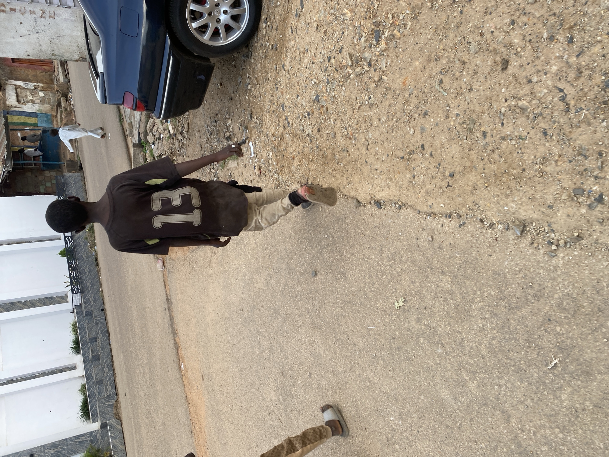 A young Black male wearing a dark t-shirt with the number "13" on the back and light-colored pants walks away from the camera on a paved road in Jos, Nigeria. He is wearing slip-on sandals. To his left, another person's legs and feet are visible, also wearing slip-on sandals. A dark blue sedan is parked on the right side of the road. In the background, there are buildings, some with signs of wear, and other pedestrians. The environment appears to be an urban street scene.