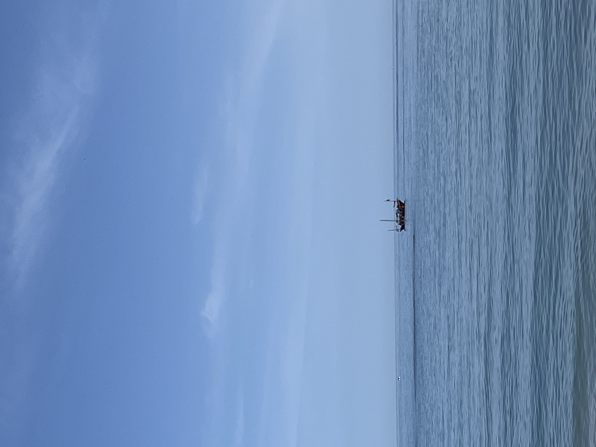 The image, presented rotated 90 degrees clockwise, depicts a serene seascape under a clear blue sky. A single, distant sailing boat, possibly a schooner, with visible masts and some rigging, floats calmly on the vast expanse of the sea. The boat is positioned near the horizon line, which sharply divides the sky from the water.
The sky, occupying the upper portion of the correctly oriented image, is a bright, expansive blue with only a few wispy, translucent clouds scattered across it. The water, filling the lower portion, appears calm with subtle, vertical ripples suggesting gentle movement or reflections. The predominant colors are various shades of blue.
The scene suggests a peaceful daytime setting, likely late morning or early afternoon, given the bright, clear conditions. The weather appears fair and tranquil, contributing to a sense of calm. This coastal vista is consistent with the provided location context of Arroyo de la Miel-Benalmádena Costa, Spain. There are no people or visible text in the image.