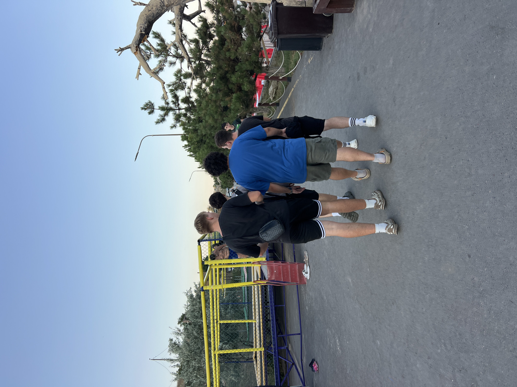 A group of four young men are walking away from the camera on a paved walkway. The man in the foreground on the left is wearing a black t-shirt, black shorts with white stripes, and a black fanny pack across his chest. He is wearing light-colored shoes and socks. The man next to him is wearing a blue t-shirt and olive green shorts, with light-colored shoes and socks. The third man is wearing a black t-shirt and black shorts, with white socks and white shoes. The fourth man, partially obscured, is wearing a black t-shirt and black shorts, with white socks and white shoes.
To the left of the walkway is a yellow and blue playground structure with a red ladder. Scattered on the ground near the structure are several pairs of sandals. In the background, there are trees and a clear blue sky. To the right of the walkway, there are more trees, a wooden railing, and a dark brown trash bin. The overall environment appears to be an outdoor recreational area. The location is specified as the city of Fontanka, Ukraine.