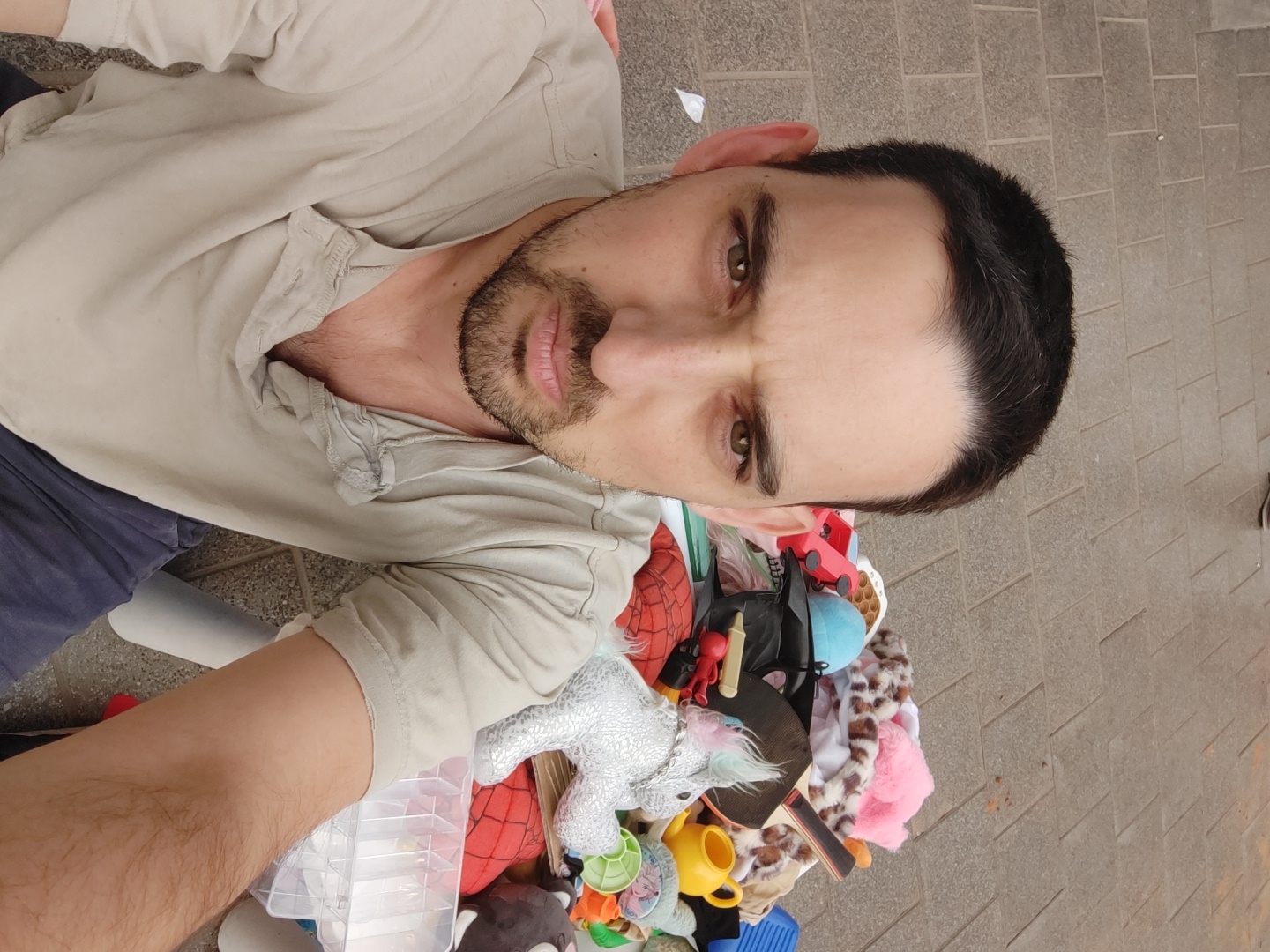 The image shows a man taking a selfie from an elevated angle, looking down at a collection of items. The man has short dark hair, brown eyes, and a beard. He is wearing a light beige shirt and dark pants.
Below him is a pile of various objects, including toys (such as a Spider-Man plush, a silver unicorn plush, a yellow watering can, and a blue toy), a table tennis paddle, and some containers.
The background appears to be a paved surface, possibly a sidewalk. Given the information provided, it is presumed to be within the city of Tel-Aviv, Israel, but without further visual details, the specific location is not possible to ascertain.