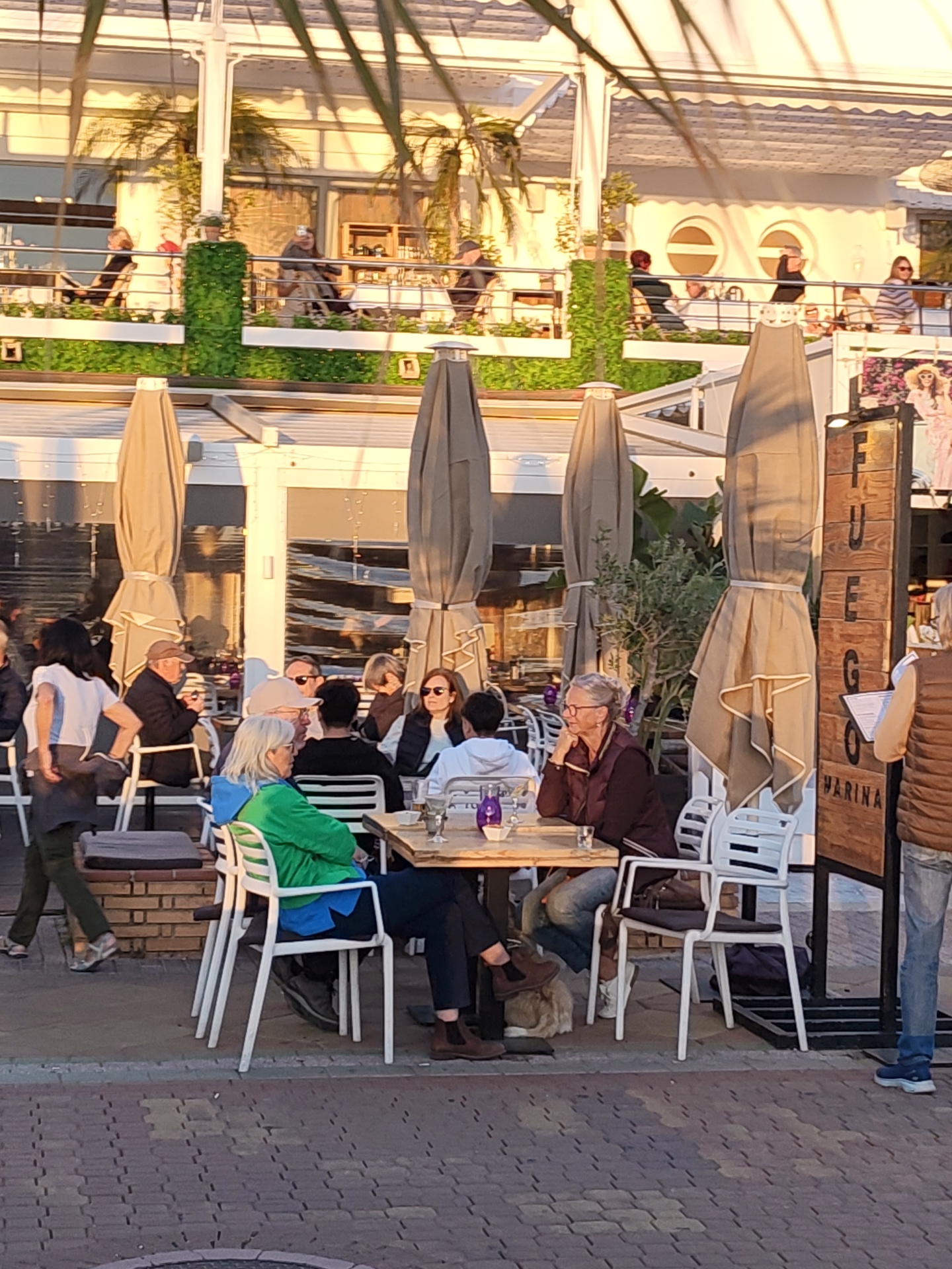 The image captures a scene at an outdoor cafe in Dénia, Spain. Several people are seated at tables under large, folded umbrellas, enjoying a meal or drinks. The cafe appears to have a second level with additional seating along a railing decorated with greenery. The architecture is predominantly white with awnings. The street is paved with patterned bricks. People are dressed in casual attire. A sign with the word "FUEGO" vertically written is visible. Palm trees frame the scene.