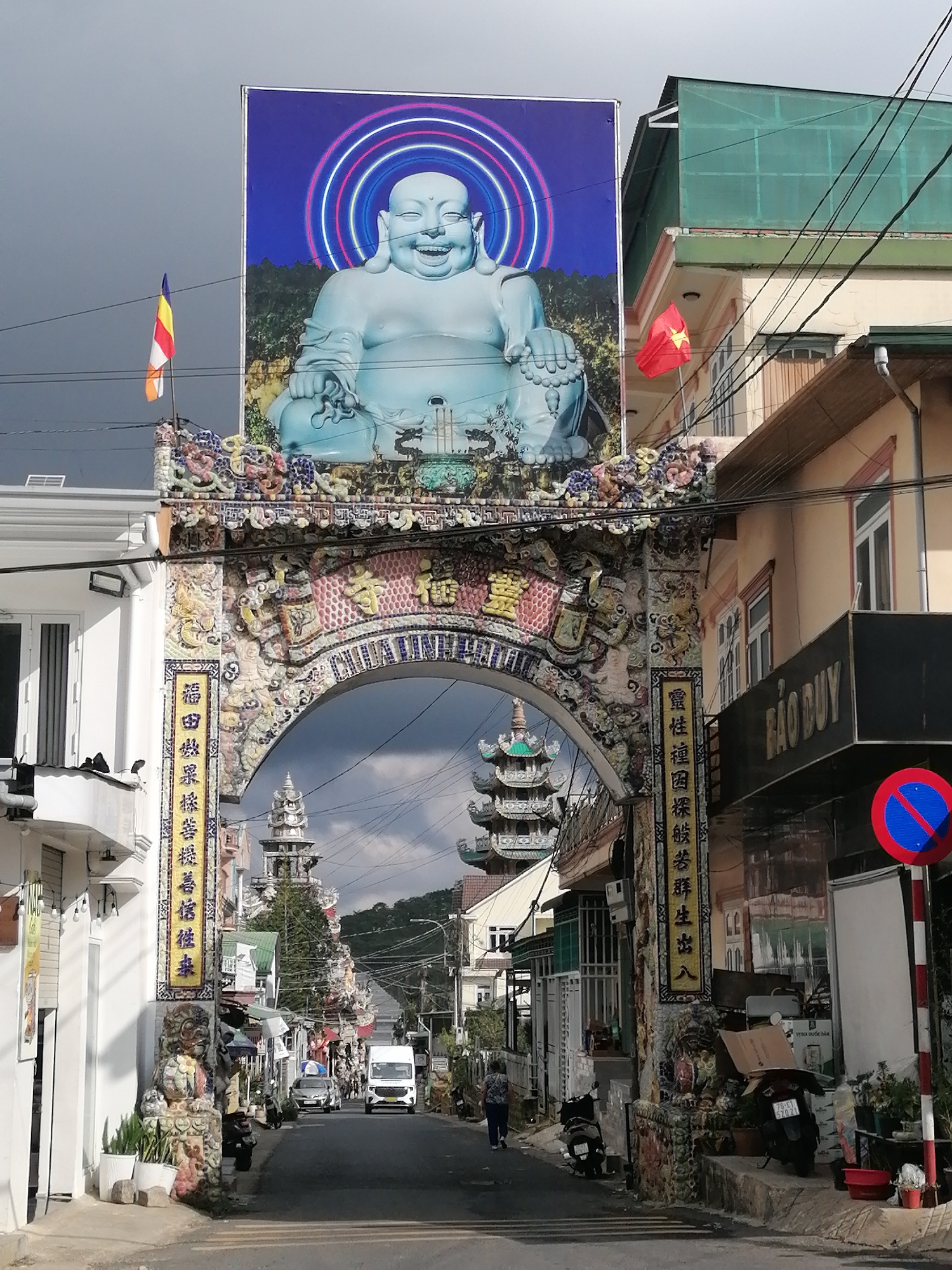 This is a full-shot, eye-level, outdoor, daytime photograph taken in Xuân Thành, Vietnam. The image depicts a street scene framed by an ornate, mosaic-covered archway. Dominating the top of the image is a large, bright billboard featuring a smiling, plump Buddha figure with neon-colored rings around its head, set against a dark, cloudy sky. To the left of the billboard, a Vietnamese flag is visible.
The archway itself is intricately decorated with colorful mosaics depicting dragons, figures, and various symbolic elements. Two vertical yellow signs with Vietnamese script are affixed to the pillars of the archway. Through the arch, a street stretches into the distance, lined with buildings of varying architectural styles, some with shops displaying signage. Several tiered pagodas or towers can be seen further down the street.
On the street, a white van is driving away from the camera, and other vehicles, including cars and motorcycles, are parked or in motion. A few individuals are visible walking on the sidewalk or in the street. The overall impression is of a lively street in a town with a strong cultural and religious influence, evidenced by the large Buddha image and the pagodas. The lighting suggests it might be an overcast day, with patches of sunlight illuminating parts of the scene.