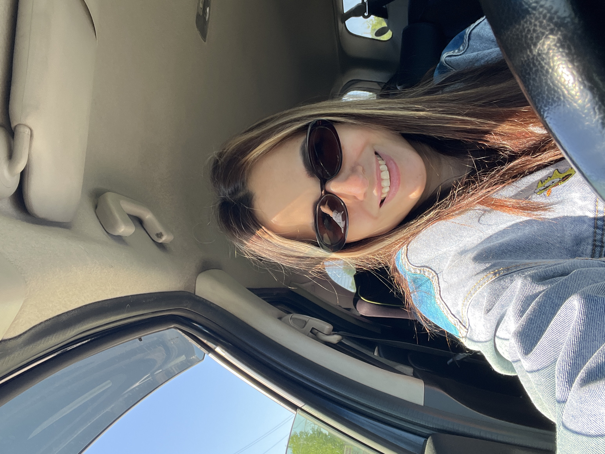 A woman with long brown hair and sunglasses is smiling at the camera from inside a car. She is wearing a denim jacket with a cartoon character on the shoulder. The interior of the car is visible, including the steering wheel, dashboard, and ceiling. Outside, a blue sky and green trees can be seen through the window. The location in the city is unknown.