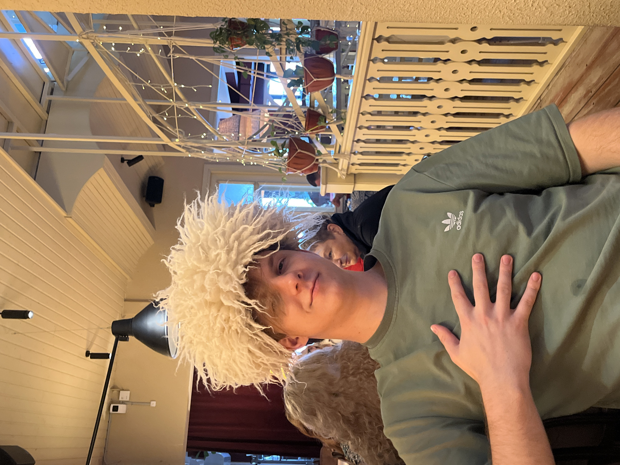 This media file shows a young person wearing a fluffy white hat. The person is smiling and has their hand placed on their chest, over a green t-shirt with an Adidas logo. The background suggests an indoor setting with white slatted railings, hanging plants, and string lights. There is another person visible behind the subject, looking towards the camera. A large lamp is also present in the scene. The image is oriented sideways, with the top of the frame appearing to the left. The lighting suggests it could be daytime, but the specific time of day and weather are not determinable. There are no clear location cues beyond it being an indoor space, possibly a cafe or a decorated room. The overall mood appears casual and playful, with the subject's smile and the novelty hat.
