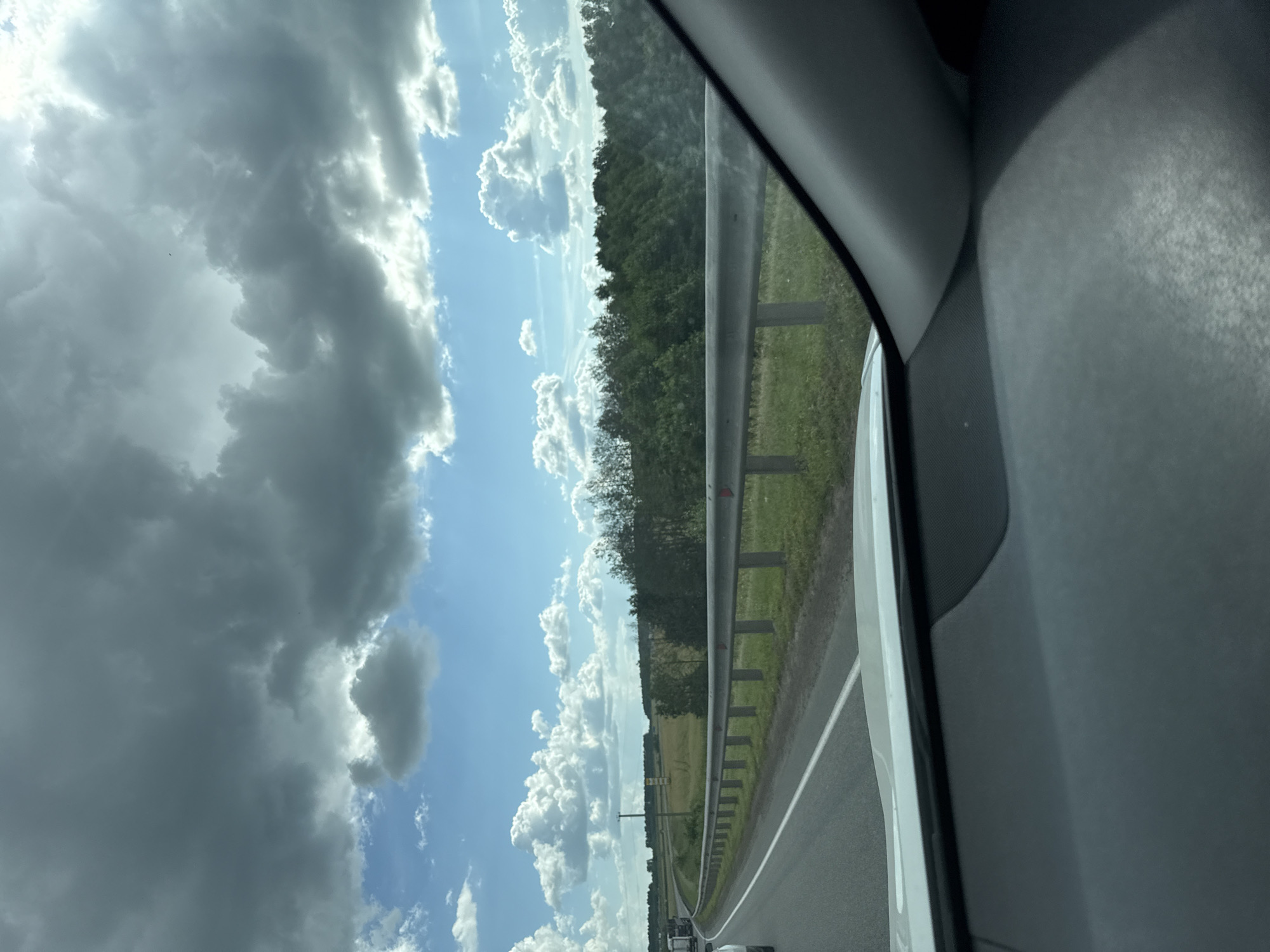 An over-the-shoulder view from a vehicle looking out at a highway on a cloudy day. The road curves to the left, with a metal guardrail running along the right side. Beyond the guardrail, a grassy field gives way to a dense line of green trees. The sky is a mix of dark, heavy clouds and patches of bright blue sky with fluffy white clouds. The interior of the vehicle is visible in the foreground, with the dashboard and windshield partially obscuring the view. There are no people visible in the frame.