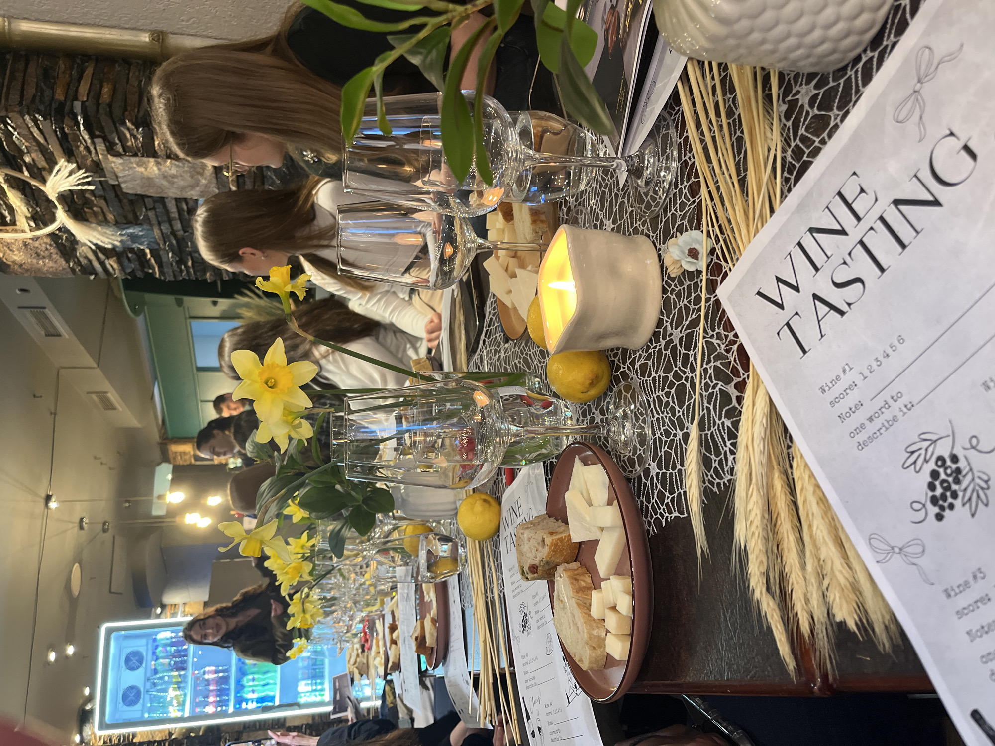 The image shows a wine tasting event taking place indoors. Several people are seated at a long table. The table is set with wine glasses, plates of bread and cheese, small lemons, and lit candles. There are also decorative elements such as yellow flowers and wheat stalks, along with "Wine Tasting" score sheets. The background shows a wall decorated with stones and a refrigerated display case. It is difficult to determine the precise location in Varna, Bulgaria from this image.