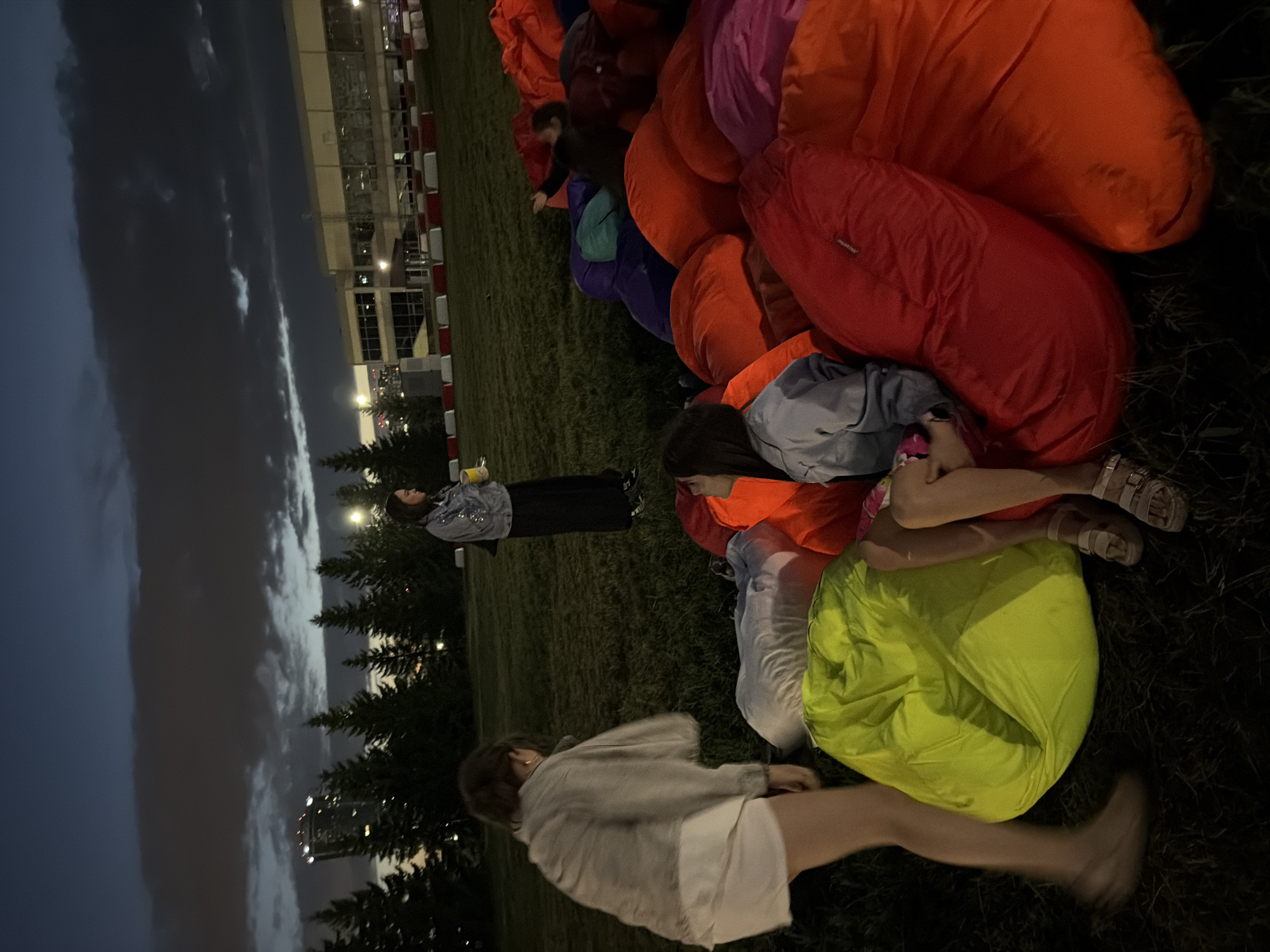 The image shows a group of people sitting or lying on a grassy area, surrounded by large, colorful beanbag chairs or cushions. The beanbags are predominantly orange, red, pink, purple, and green. The scene appears to be outdoors, with trees and a building in the background. The sky is dark and cloudy, suggesting it is either dusk or nighttime. Lights from the building and potentially streetlights are visible.
Several individuals are present. In the foreground, a person is walking away from the camera, their legs blurred due to motion. To the right, another person is lying down on a lime green beanbag, with their legs visible. Further back, a group of people are interacting with the beanbags. One person is seated, leaning against a large orange beanbag, while another is lying down with their legs up. In the middle ground, two individuals are visible: one standing and looking towards the left, wearing a denim jacket, and another person to their left, also standing. A person in a dark outfit is seen further back, reaching towards the beanbags.
The overall mood seems relaxed and informal, possibly a gathering or an outdoor event. The time of day is clearly evening or night, given the dark sky and artificial lighting. The presence of beanbags suggests a casual setting for seating and lounging. There is no visible text in the image.