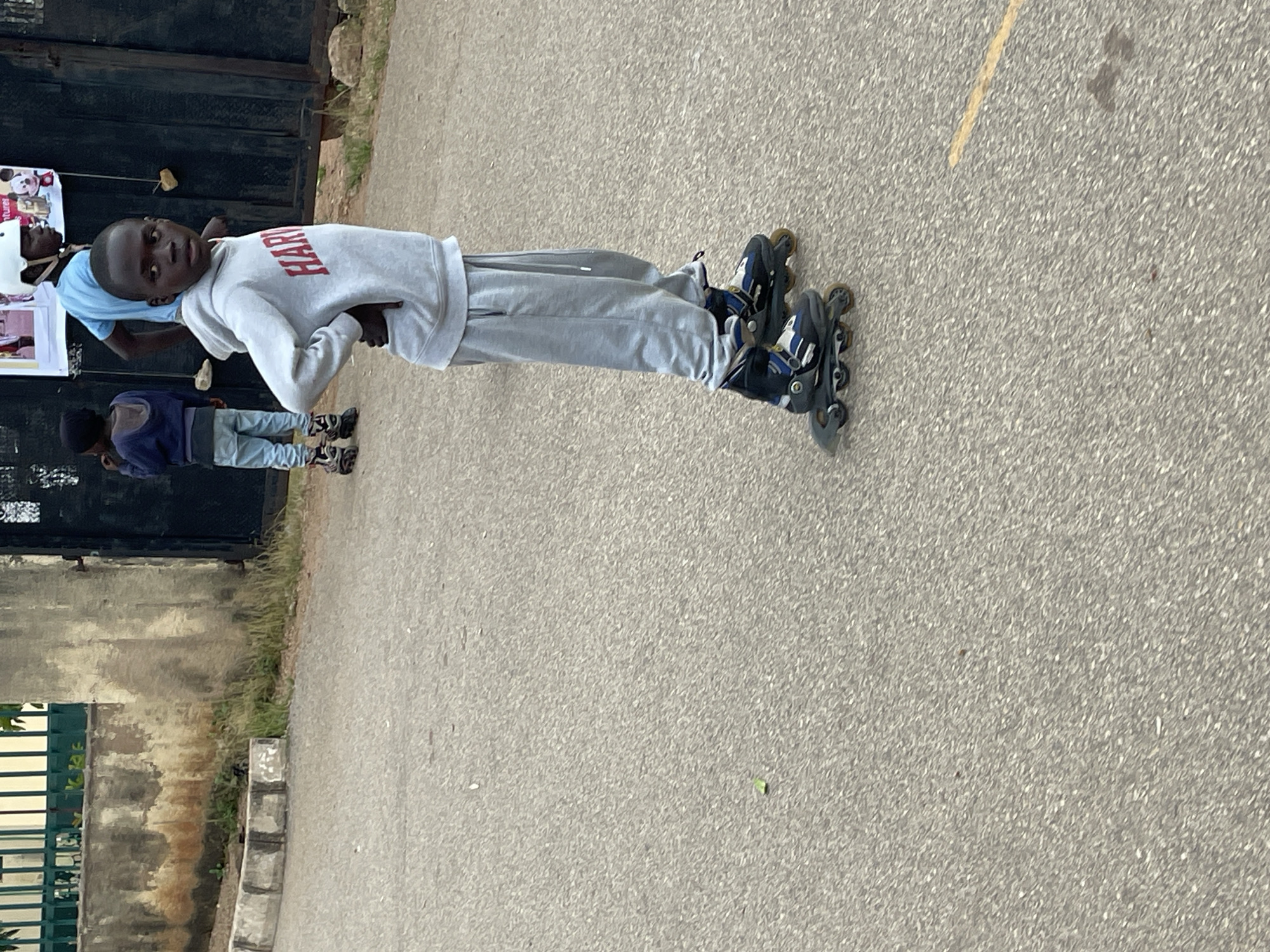In Jos, Nigeria, a young Black boy wearing a grey hooded sweatshirt with "HAR" visible on the front and grey sweatpants is shown in profile, standing on roller skates. His left hand is on his hip. Behind him, another Black boy, also wearing a blue t-shirt and a white helmet, stands near a dark gate and a poster. To the far left, a third Black boy in a blue hooded jacket and light-colored pants is visible. They are on a paved surface, possibly a road or courtyard, with a weathered stone wall and green fencing in the background.