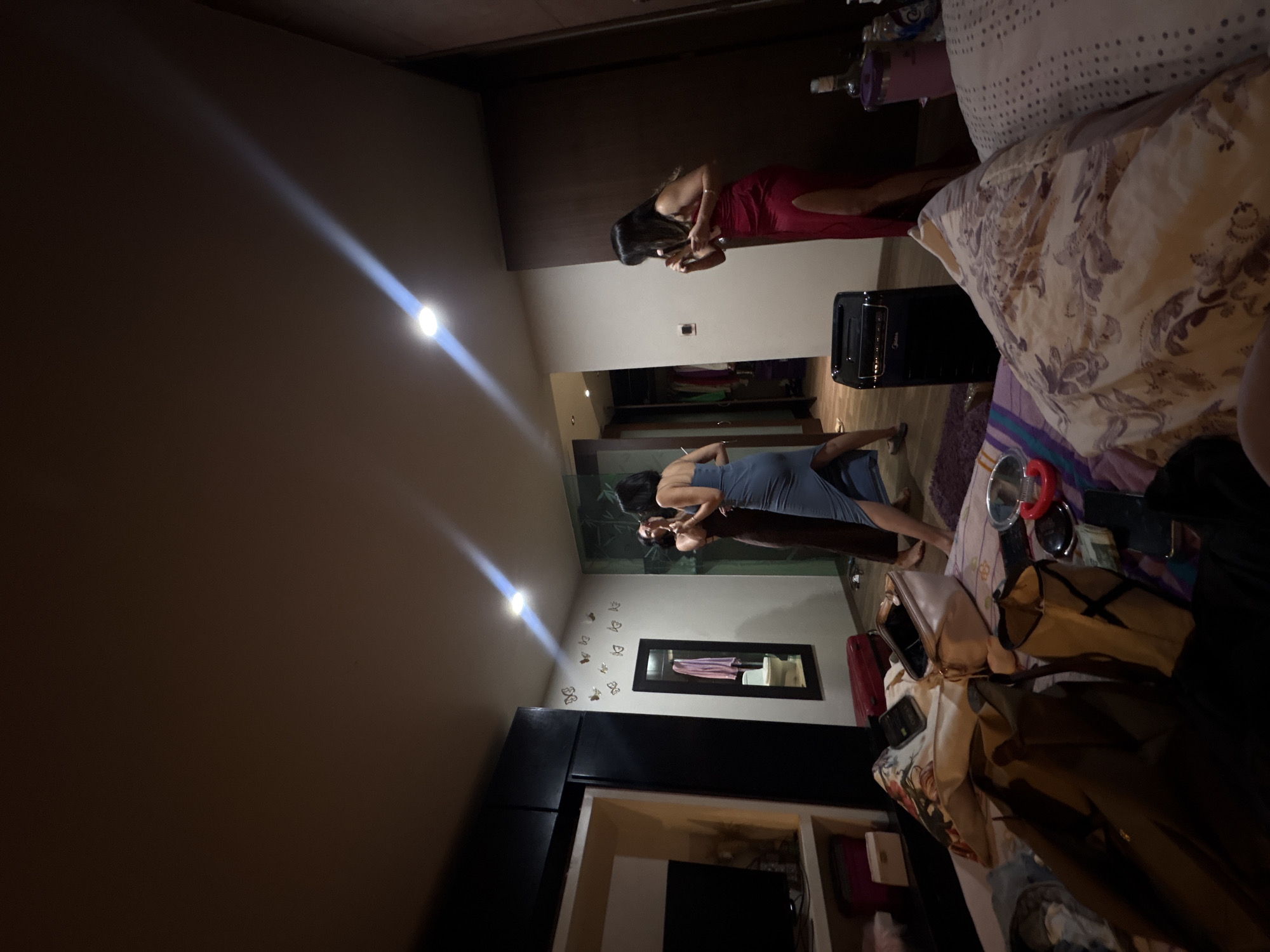 The image depicts an indoor scene, likely a bedroom, located in San Andrés Cholula, Mexico. Four women are present, three of whom are the central focus. Two women in the middle of the frame, one in a brown dress and the other in a blue dress, are engaged in a playful interaction, with their arms around each other. A third woman, in a red dress with a high slit, stands to the right, holding a glass and appearing to converse. The room is furnished with a bed, clothing items scattered on it, a mirror on the wall adorned with butterfly decals, and a dark wardrobe on the left. The lighting suggests it is nighttime or a dimly lit interior space, with overhead lights creating prominent beams. A red suitcase is visible near the mirror, and a black electronic device, possibly a shredder, is positioned between the women. The overall impression is of a casual gathering or a pre-party scene within a private residence.