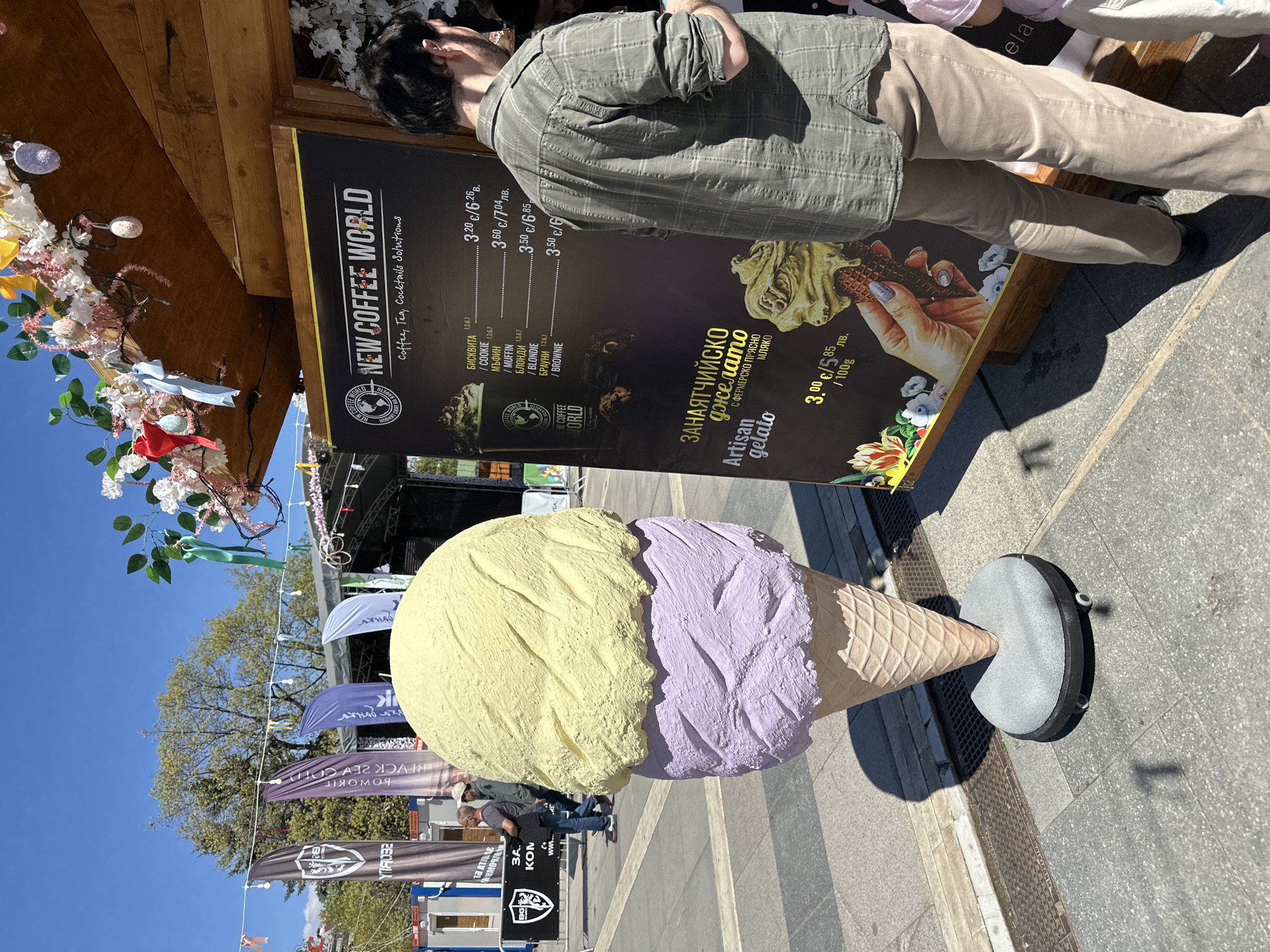 The image shows an outdoor scene in Sofia, Bulgaria. A man is looking at a menu board for "New Coffee World," which offers coffee, tea, cocktails, and desserts like cookies, muffins, blondies, and brownies, along with "Artisan gelato." In the foreground is a large, decorative ice cream cone with a yellow scoop on top and a purple scoop below. Flags with various logos are in the background. People are walking in the area near the flags. The sky is blue.