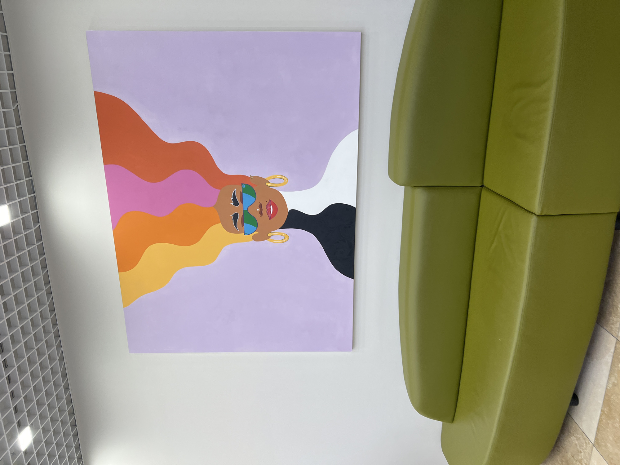 The image depicts a contemporary interior scene, likely a waiting area or lounge space. The primary subject is a colorful, abstract painting of a person's head and flowing hair against a pale lavender background. The person in the painting is depicted with sunglasses, bright red lips, and gold hoop earrings. Their hair is stylized with waves of orange, pink, and yellow, contrasting with a black shape on the right side of the composition.
To the right of the painting, a section of a modern, modular sofa is visible. The sofa is a vibrant lime green and appears to be made of cushioned sections, suggesting a comfortable and stylish seating arrangement. The texture of the upholstery is smooth and slightly reflective.
In the upper left corner of the image, a grid-like ceiling structure is partially visible. This detail, along with the plain white walls, contributes to a clean and minimalist aesthetic. The lighting appears to be artificial, casting subtle highlights on the sofa and ceiling.
The overall mood is cheerful and modern, with the bold colors of the artwork and the sofa creating a focal point. There are no visible signs of a specific time of day or weather conditions, but the indoor setting implies controlled environmental conditions. The location is identified as Yekaterinburg, Russia. No text is visible in the image.