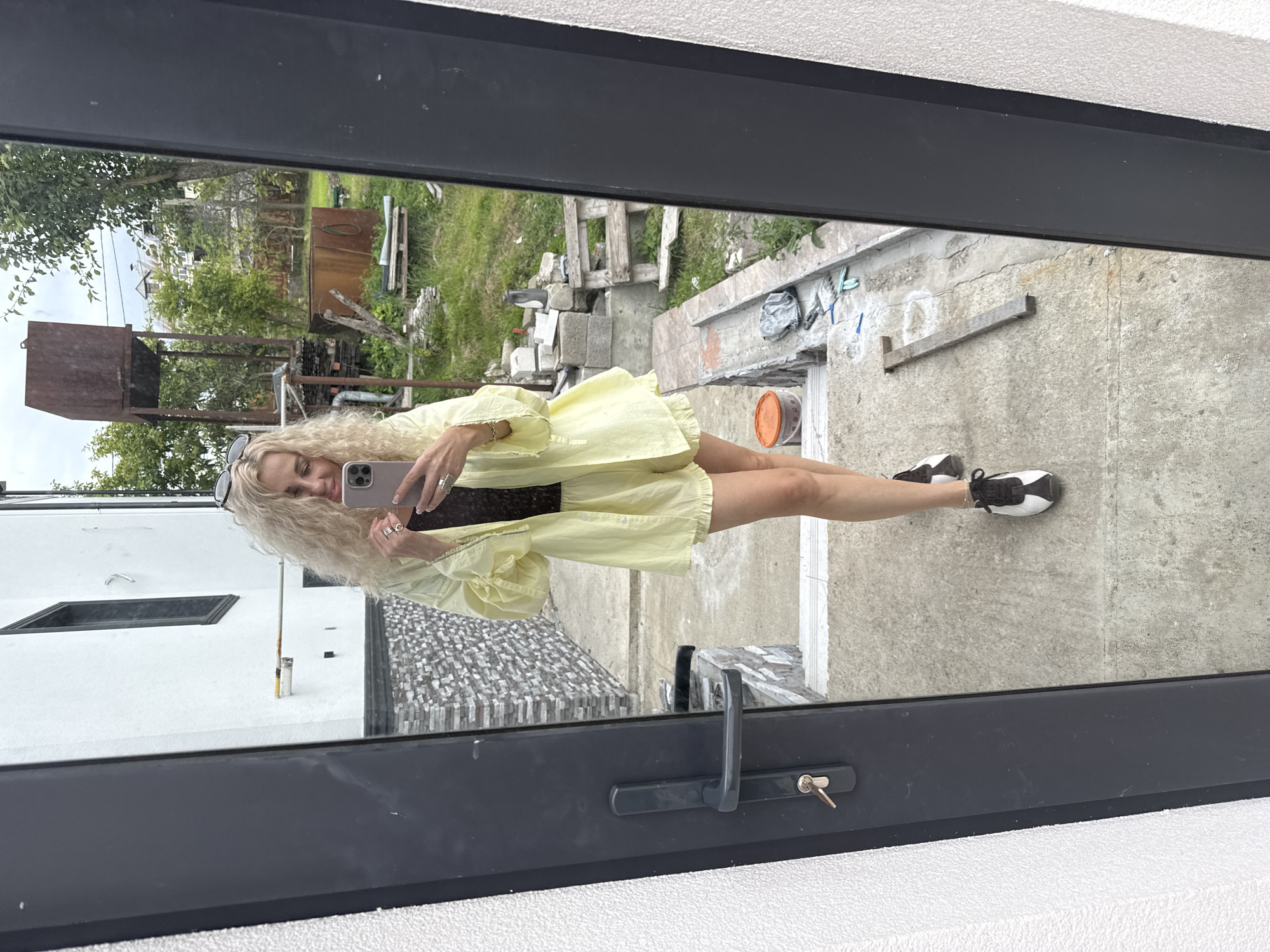 A woman with curly blonde hair is taking a selfie in a mirror. She is wearing a light yellow ruffled shorts set and black and white sneakers. She is standing on a concrete pathway outside a white building with gray stone accents. In the background, there is a yard with trees, construction materials, and a rusty water tower. The scene is located in Baranovka, Russia.