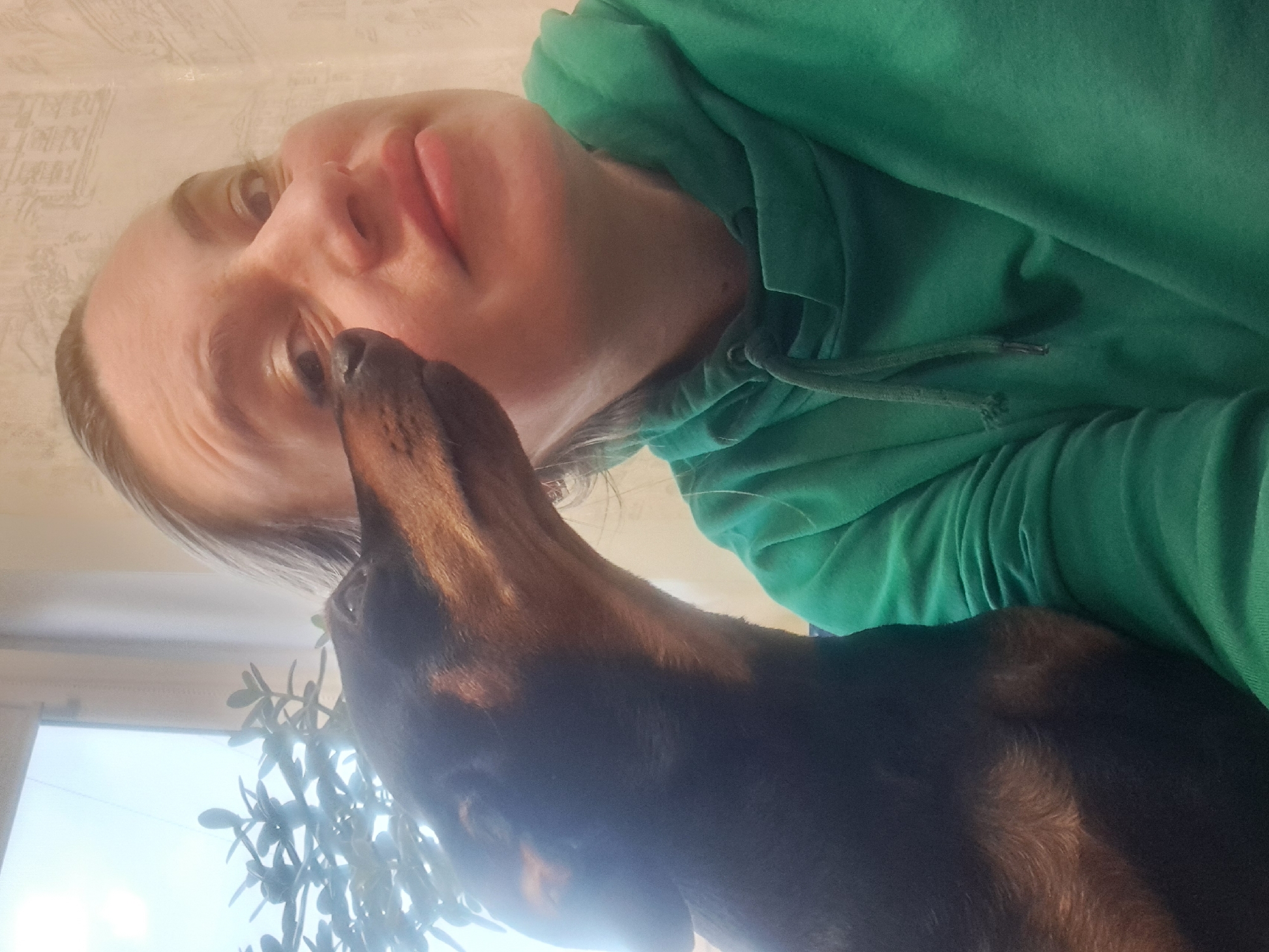 A low-angle shot shows a woman with fair skin, light hair, and a green hooded sweatshirt looking down at a dachshund. The dog, mostly black with brown on its head and neck, is facing the woman. Behind the dog, partially obscured, is a houseplant and a window revealing bright outdoor light. The wallpaper on the wall behind the woman is light-colored with a subtle pattern. The location may be Elektrostal, Russia.