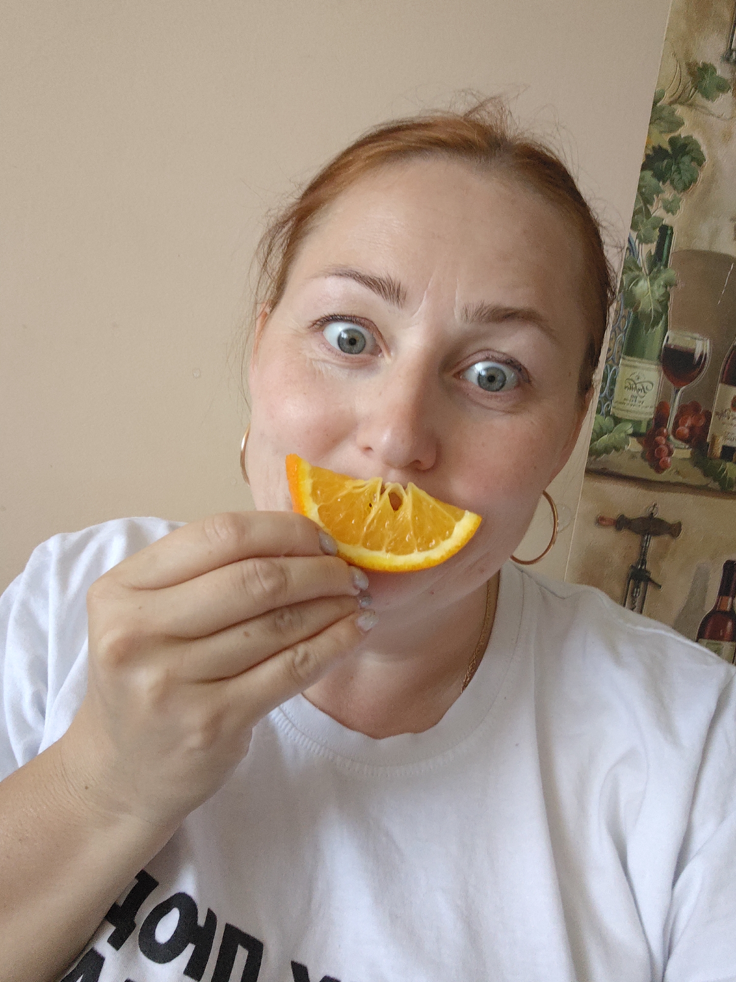 A medium shot depicts a fair-skinned woman holding a slice of orange against her mouth. She has reddish-brown hair pulled back from her face, light blue eyes, and is wearing gold hoop earrings and a white t-shirt. The shirt has some indiscernible dark-colored text on it. Her fingernails are painted with a light color, and she is wearing a gold chain necklace. Behind her, to the right, is a wall with a wine-themed graphic featuring bottles and glasses of wine, grapes, and a corkscrew. The rest of the background is a plain, light-colored wall. The location given is Марковский, Russia, however, this is not verifiable within the image.