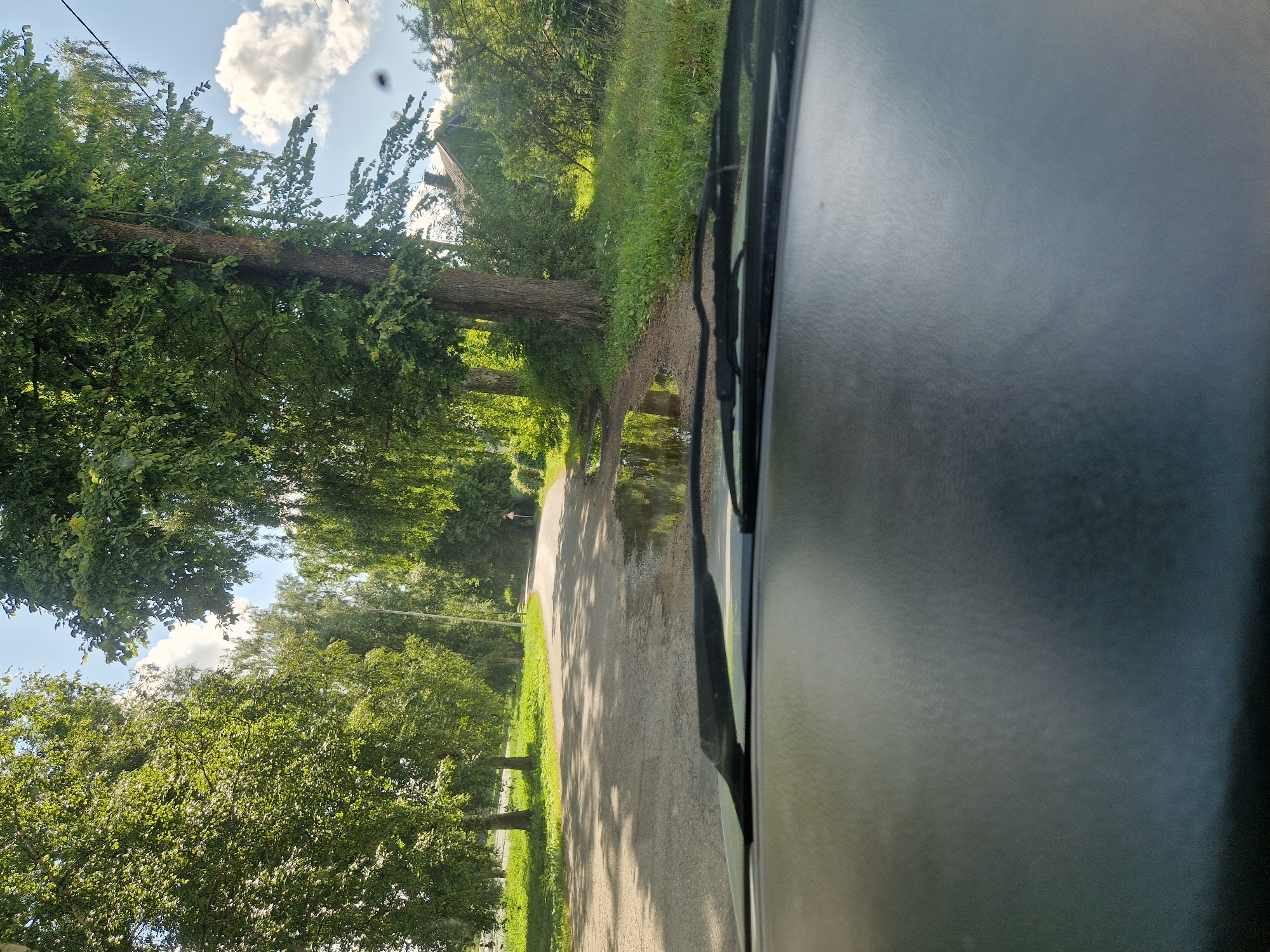 A driver's perspective shot from inside a car shows a rural road lined with trees. The road is paved and has puddles from recent rain. On the right side of the road, there is a wooden house with a dark roof, partially obscured by dense vegetation. A traffic sign is visible on the right side of the road. The sky is blue with scattered white clouds. The foreground of the image is dominated by the dashboard of the car. The location is identified as Radopole, Latvia.