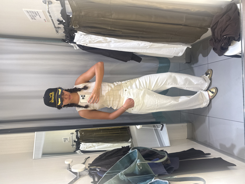 The scene depicts the interior of a retail clothing store fitting room, located in Los Angeles, United States of America.
A single individual, an adult female with dark hair styled in two braids, is centrally positioned, facing a mirror and holding a light-colored smartphone in her right hand, appearing to capture a self-portrait. She is wearing a black baseball cap with yellow embroidery, a light-colored ribbed sleeveless top, white wide-leg jeans with cuffed hems, and gold-colored slip-on shoes with small tassels. A gold-toned bracelet adorns her left wrist, and her left hand is placed within the front pocket of her jeans.
The fitting room walls are light grey, and the floor is composed of light grey tiles. A large rectangular mirror is mounted on the left wall, reflecting parts of the room, including a partially visible white stool and additional clothing on hangers. To the right, a grey privacy curtain with a light blue/teal bottom trim is closed. A metal rail extends from the right wall, supporting several garments on black hangers, including dark items, a pair of white pants, and a pair of dark olive-colored wide-leg pants. A white circular stool in the lower right foreground holds a folded dark brown garment. A white sign with black text and a red rectangular base is affixed to the upper right wall. The lighting appears to be artificial overhead illumination.