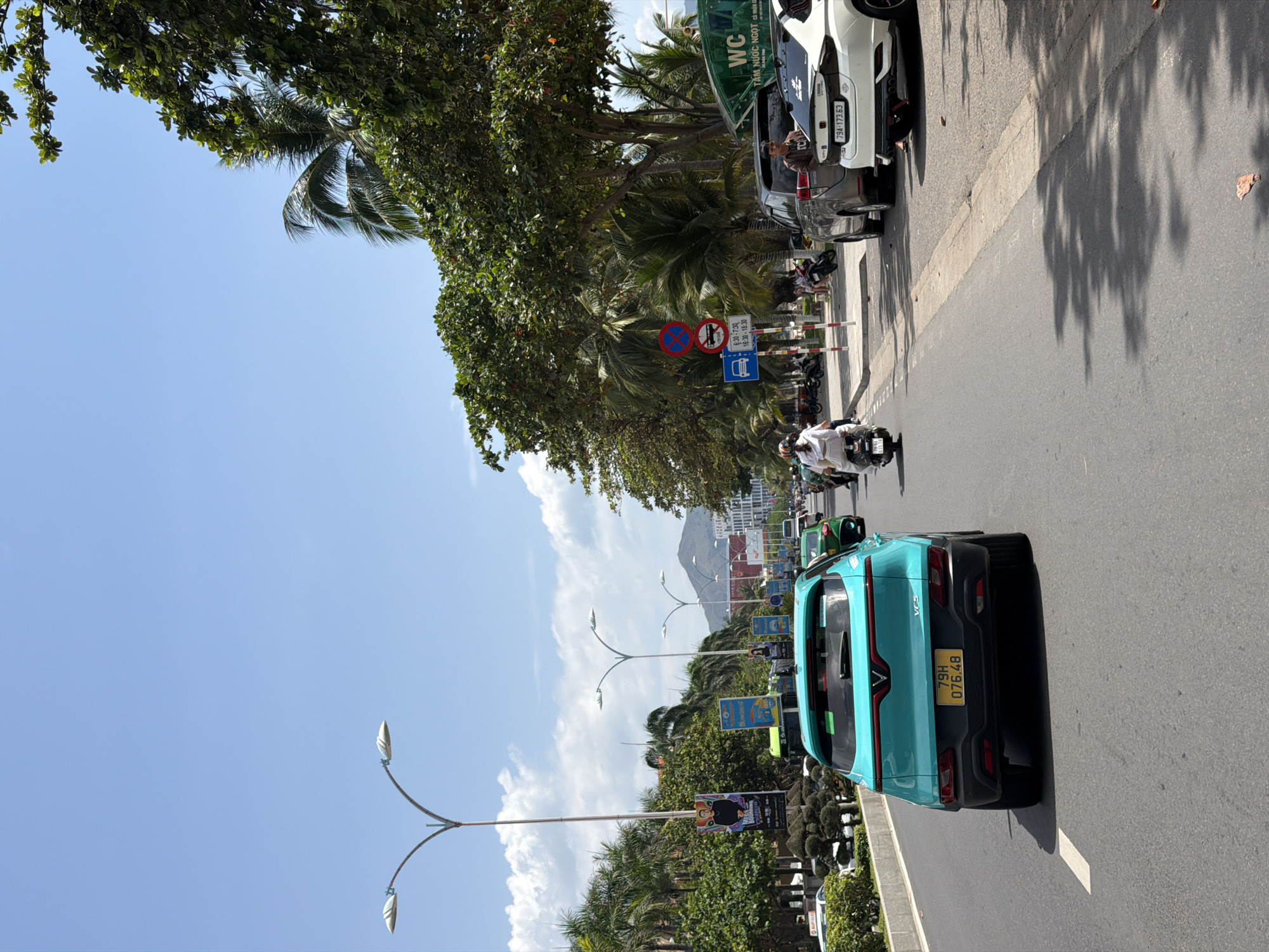 The image captures a sunny daytime street scene in Nha Trang, Vietnam. A bright teal VinFast VF 5 electric car, bearing license plate "79H 076.48", is prominently featured on the right, driving on an asphalt road. To its left, a tree-lined sidewalk separates the road from a row of parked vehicles, including a white car with license plate "79A-173.63" and a grey SUV. Several people are visible, riding motorbikes or sitting in parked cars.
The setting is an urban environment with lush greenery, including large trees with dense foliage and tall palm trees. In the distance, mountains rise under a clear blue sky with scattered white clouds. Modern streetlights with distinctive twin-lamp designs illuminate the area.
Various road signs are visible: a red circular sign indicating "no entry" or "no parking," another red circular sign prohibiting trucks and motorbikes, a blue rectangular parking sign with specific time restrictions ("6:30-7:30" and "16:30-18:30"), and a blue sign with a bus symbol, possibly indicating a bus stop. A green sign attached to a parked vehicle reads "WC" (toilet) and "TẤM NƯỚC NGỌT CÓ NHẬN GI". Advertising banners are strung along the street, with one displaying a person's face. The bright sunlight casts distinct shadows of trees and vehicles on the road, suggesting midday or early afternoon.