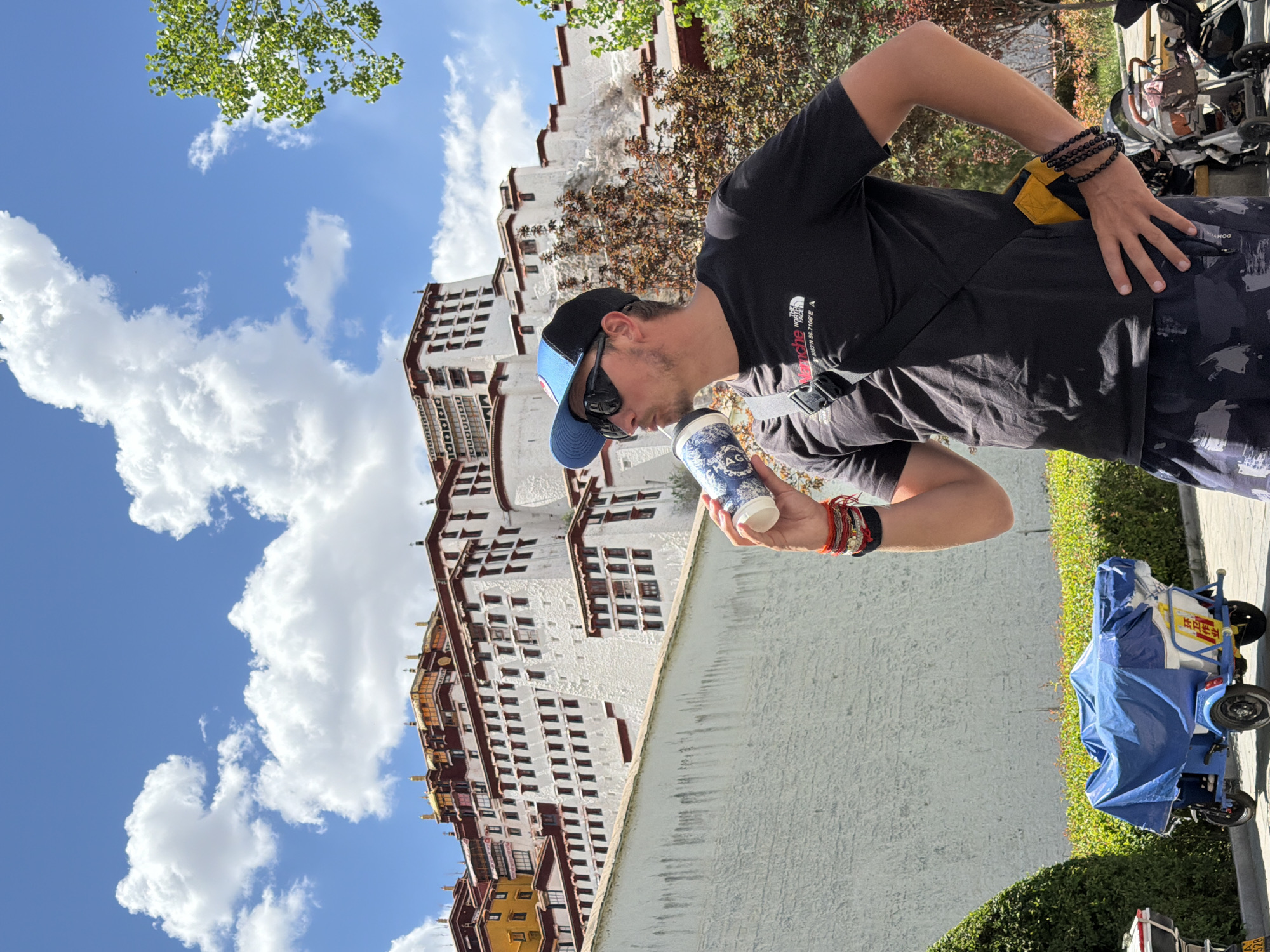This image captures a man standing outdoors on a bright, sunny day, with the majestic Potala Palace forming a significant backdrop. The man, positioned slightly to the right of the center, is wearing a black short-sleeved t-shirt, dark patterned shorts, a blue and black baseball cap, and sunglasses. He is holding a white and blue patterned cup, seemingly taking a drink or inspecting its contents. On his left wrist, he wears multiple bracelets, including beaded ones and colorful string bands. His right hand is placed on his hip. His black t-shirt features the "THE NORTH FACE" logo and coordinates "N 27°59'36.918" E 86°55'37.710", which point to Mount Everest. The cup he holds displays the letters "AG".
Behind the man, the iconic white facade of the Potala Palace, characterized by its numerous windows and traditional Tibetan architectural style, rises impressively under a clear blue sky dotted with fluffy white clouds. Green foliage from trees is visible in the upper left, and some brownish shrubs add texture near the palace walls. To the far right, partially visible, is a small, blue-covered tricycle or cart with yellow accents, on which some indistinct Chinese characters are present. The strong sunlight indicates it is daytime, likely late morning or early afternoon. The image provides a glimpse of a tourist enjoying a refreshment in front of a significant cultural landmark in Chengguan District, China.