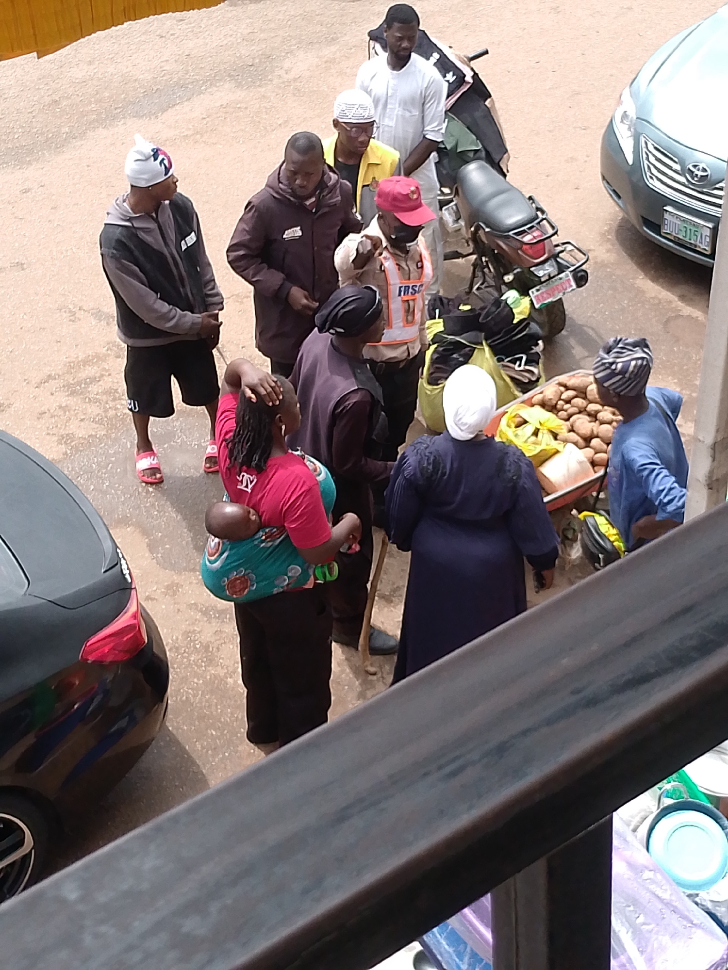 In Bukuru, Nigeria, a group of people are gathered near a road. A black car is parked on the right side of the frame, with its front bumper and hood visible. A motorcycle is parked behind the car. In the foreground, a woman is carrying a baby wrapped in a colorful cloth. She is wearing a red t-shirt and dark pants. Another woman, wearing a dark blue dress and a white headscarf, is standing next to a cart filled with potatoes. A man in a blue shirt is standing beside the potato cart. A man in a red cap and an orange vest with "FRSC" written on it is visible in the middle of the frame. Several other individuals, including men and women, are also present in the scene, engaged in various activities. The ground appears to be paved with asphalt. The overall scene suggests a marketplace or a busy street in a Nigerian city.