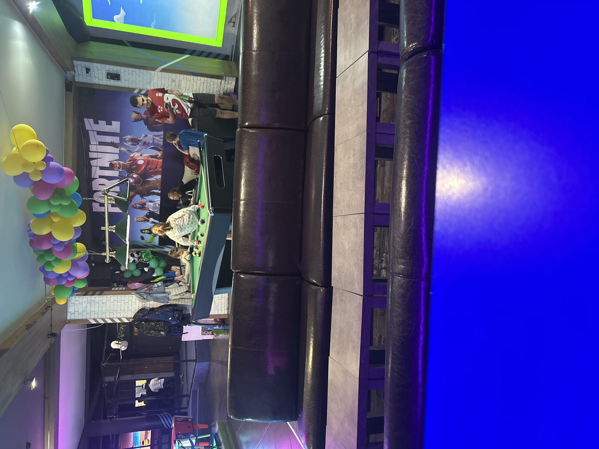 A brightly lit entertainment venue features a pool table surrounded by patrons. A "Fortnite" themed backdrop with a large screen above it adorns the wall. A column of colorful balloons is visible on the left. Nearby, a tiered play structure or seating area is evident. The venue appears to be located in Burgas, Bulgaria.