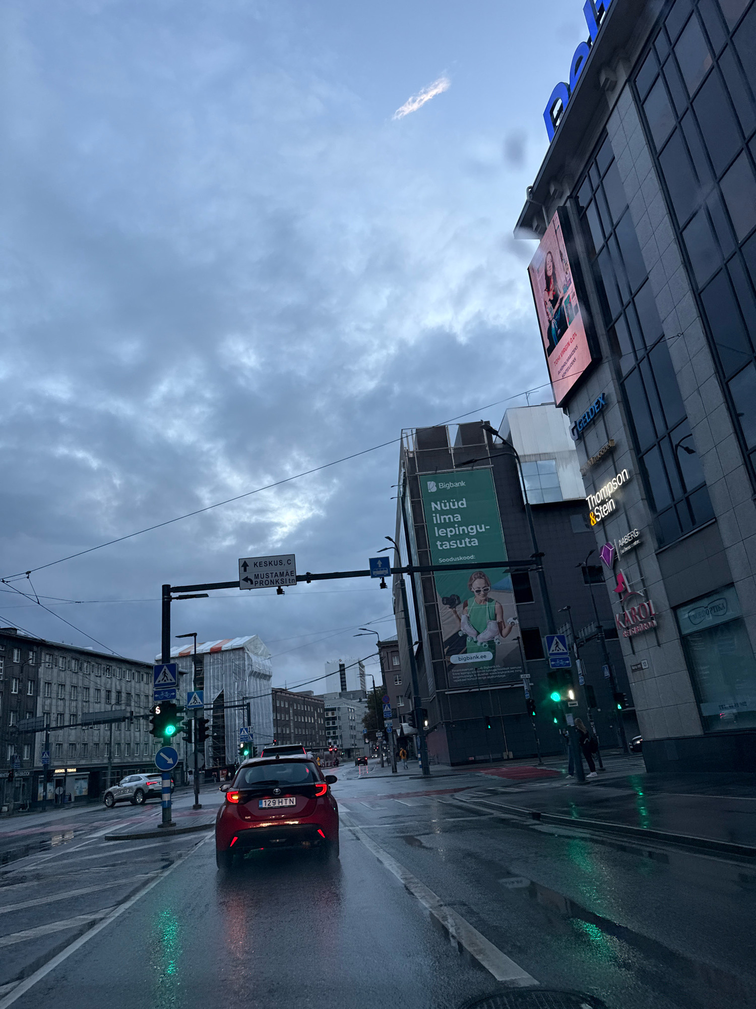 The image captures a street scene in Tallinn, Estonia, under a cloudy, overcast sky, suggesting a potentially rainy or recently rained-on day, as evidenced by the wet road surfaces reflecting the streetlights and traffic signals. In the foreground, a red car with license plate "129 HTN" is at a traffic light, which is currently green. The car appears to be a Toyota Yaris.
To the right, a modern building with large glass windows is visible, displaying various business names such as "Bigbank," "Geldex," "Thompson & Stein," "NABERG," and "KAROL." A large advertisement for "Bigbank" featuring a woman on roller skates is prominently displayed on a billboard attached to another building.
In the background, a row of older, gray-toned apartment buildings lines the left side of the street. Traffic signals and street signs are visible throughout the scene, indicating a busy urban intersection. One street sign indicates directions to "KESKUS, C," "MUSTAMÄE," and "PRONKSI tn." The presence of traffic lights, street signs with Estonian text, and the architectural styles collectively point to a location within the city of Tallinn. People are seen walking on the sidewalk and crossing the street, suggesting daily urban life. The overall atmosphere is one of a typical city environment, possibly during the late afternoon or early evening, given the subdued lighting.