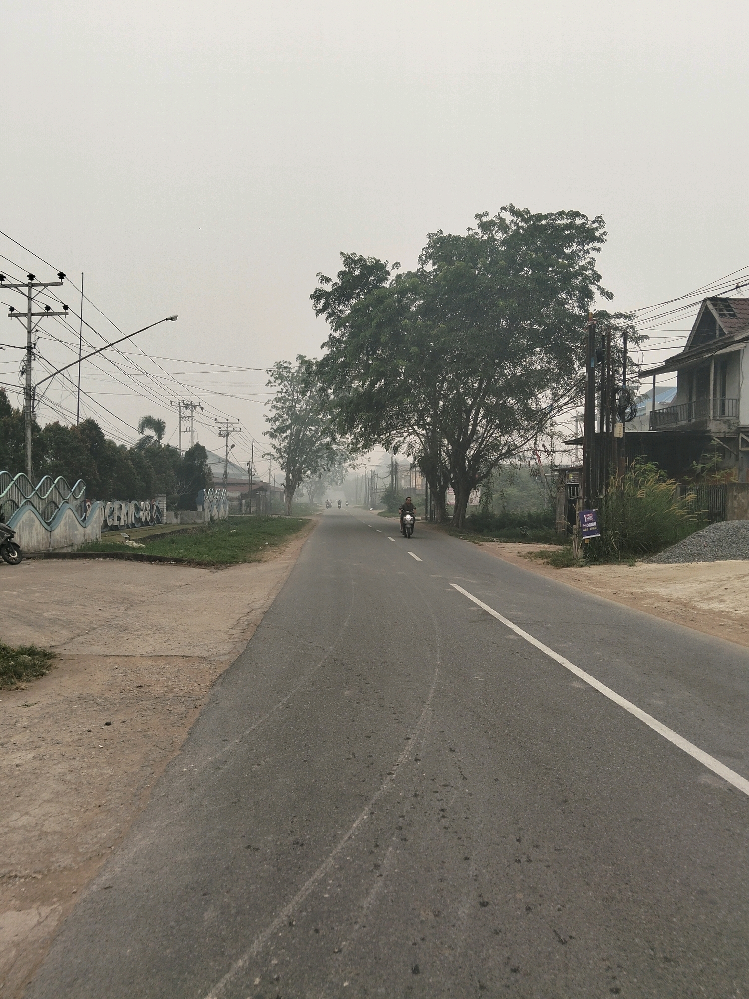 The image depicts a road in Sungai Raya, Indonesia, on a hazy day. The asphalt road, with a white line marking the center, stretches into the distance, curving slightly to the right. On the left side of the road, a concrete sidewalk borders a grassy area. A decorative concrete barrier with a wave-like pattern runs along the sidewalk, with some graffiti visible on it. A motorcycle is parked on the sidewalk to the far left. Beyond the barrier, there are trees and buildings.
To the right of the road, there is a patch of grass and then a dirt area with a pile of gravel. A two-story building with a weathered exterior stands prominently on the right. Several utility poles with a complex network of wires are present throughout the scene, particularly on the left side and behind the building on the right. Trees, some with dense foliage, are scattered along both sides of the road and further into the background, contributing to the hazy atmosphere. A few people on motorcycles are visible in the distance, traveling along the road. The sky is overcast and a light gray, suggesting diffused lighting conditions.