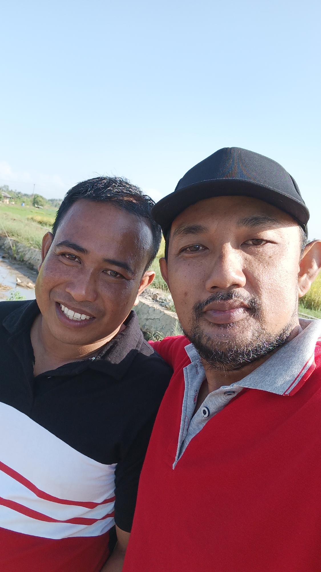 Two men are posing for a selfie outdoors, likely in the rural outskirts of Batarang, Indonesia, given the presence of a muddy ditch, lush green fields, and what appears to be a dirt path or embankment in the background under a clear blue sky. The man on the left, wearing a black polo shirt with a red and white diagonal stripe design, smiles broadly, exposing his teeth. The man on the right, wearing a red polo shirt and a black baseball cap, has a more subdued expression and sports a beard. The overall lighting suggests it is daytime, possibly late afternoon due to the warm light. The men appear to be friends or acquaintances enjoying a casual moment together in what seems to be a natural or agricultural setting.