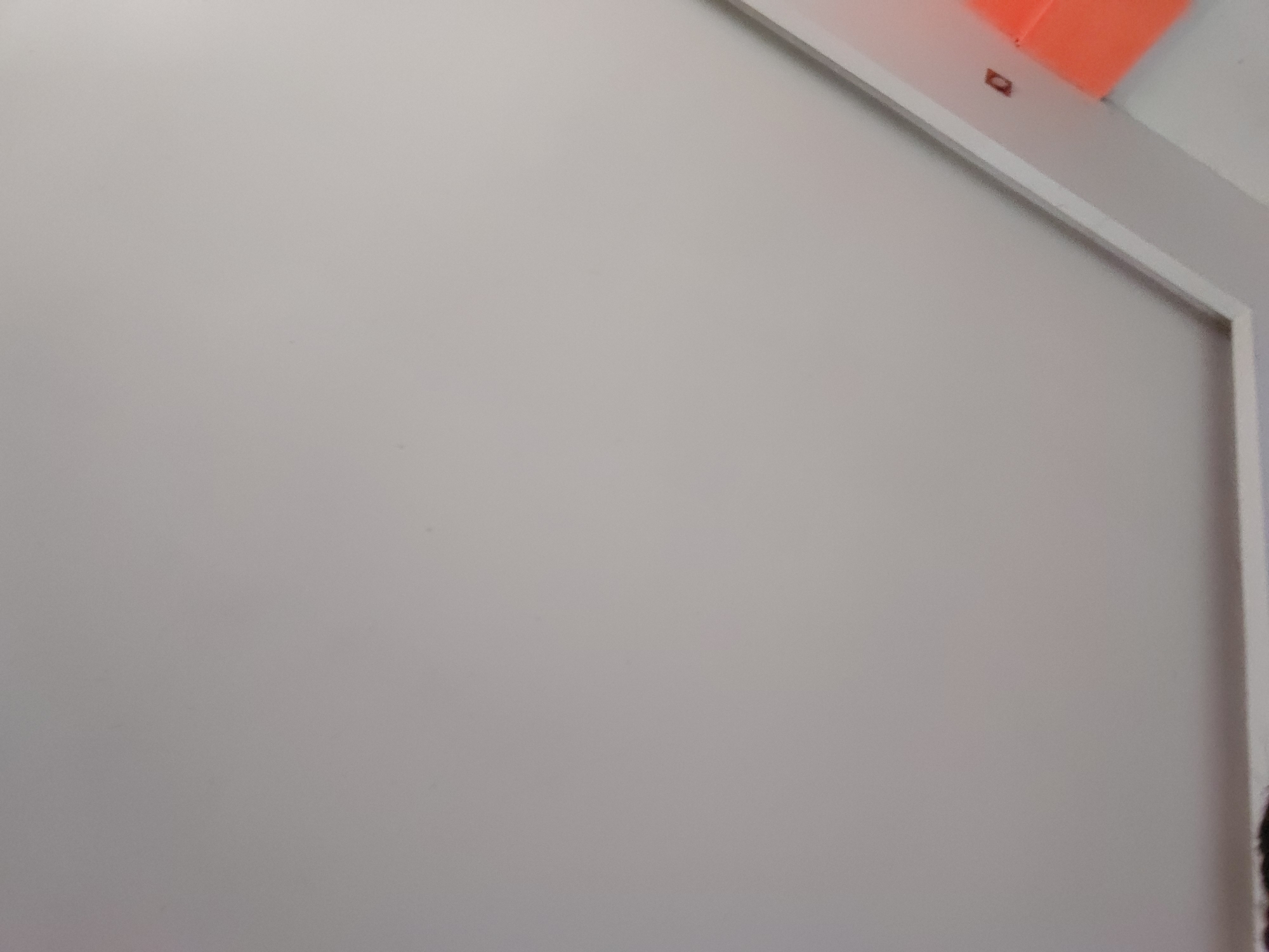 The image displays a close-up view of a plain, light grey or off-white interior wall, filling most of the frame. The wall surface appears smooth and clean.
In the top right corner, a section of a bright orange object is visible, likely a corner or edge of a piece of furniture, a decorative element, or a part of the room's architecture. Below this orange object, a crisp white trim or frame runs diagonally downwards from the top right, then vertically along the right edge of the image. This trim suggests the boundary of a wall, doorway, or window.
Affixed to the white trim, directly beneath the orange object, is a tiny, dark rectangular item. A faint, indistinct mark or symbol is barely discernible on its surface, but it is not legible text. At the very bottom right edge of the frame, a small, dark, textured area, possibly a glimpse of hair or fabric, is just visible.
The lighting is even and soft, indicating an indoor setting, likely under ambient artificial light or indirect natural light. No people are visible, and no specific activity or event is taking place. The overall scene suggests a static snapshot of an interior space, possibly located in Santa Ana, Venezuela.