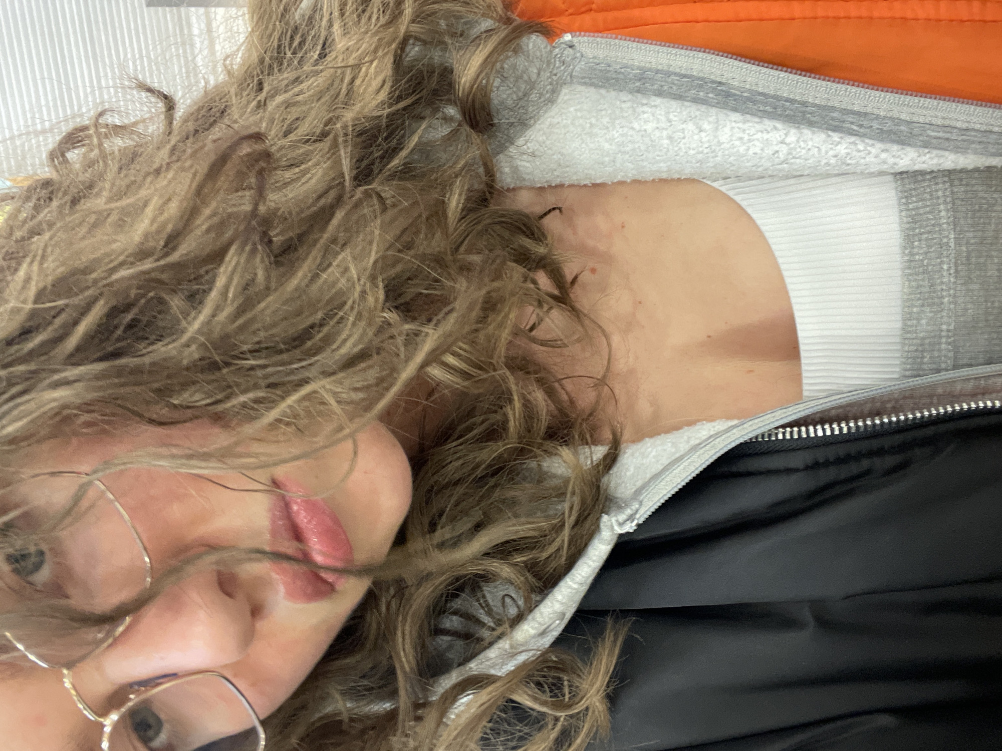 A close-up selfie shows a person with curly blonde-brown hair wearing glasses. The person's face is partially obscured by their hair and glasses. The person is wearing a white ribbed tank top, a grey fleece-lined jacket, and a black jacket with an orange accent. The image does not contain any information about the location of Moscow, Russia.
