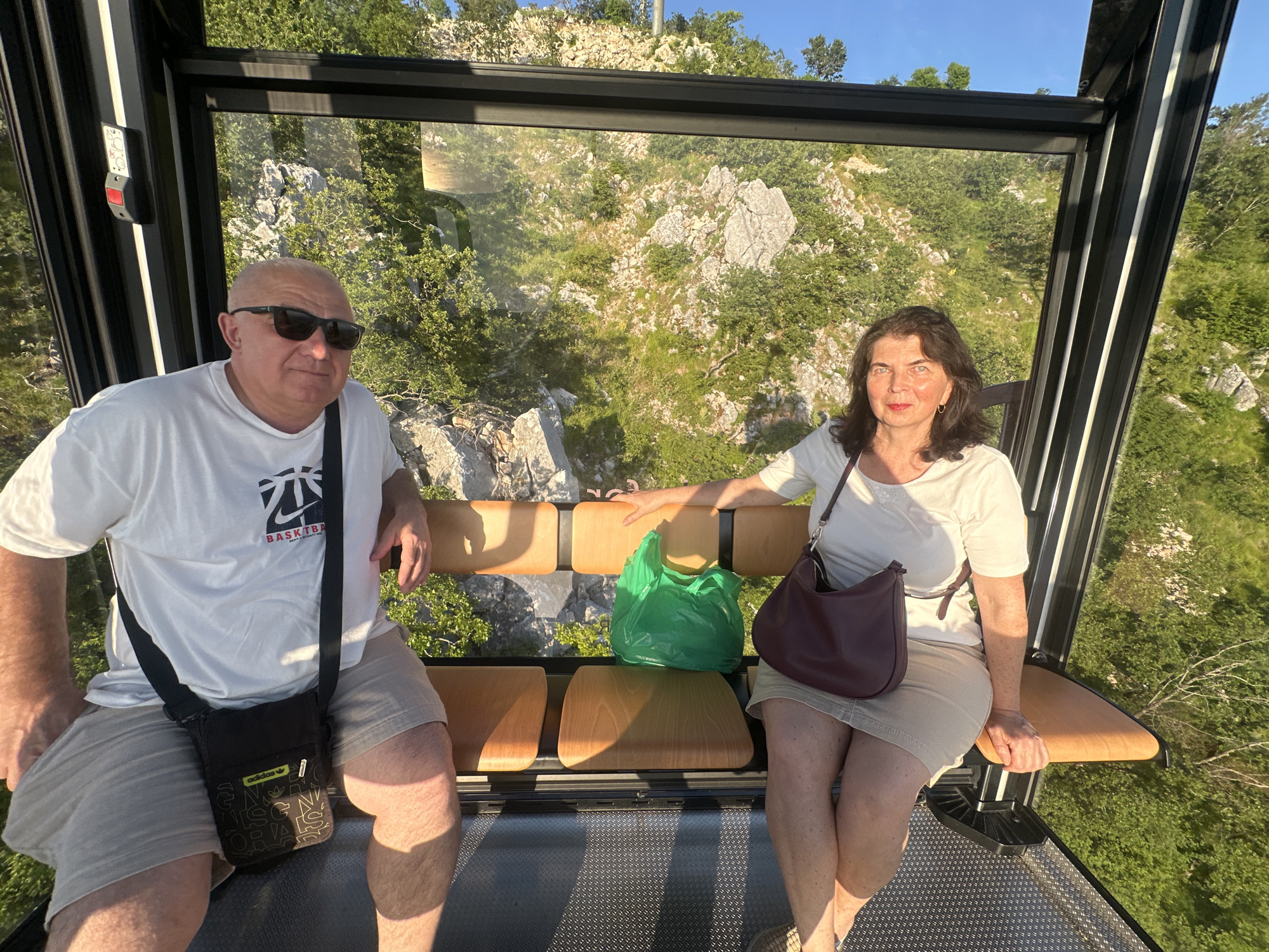 A man and a woman are seated inside a cable car with a view of the mountainous landscape of Škaljari, Montenegro. The man, on the left, wears a white t-shirt, sunglasses, and khaki shorts, with a black messenger bag slung across his chest. The woman, on the right, is dressed in a white t-shirt and a beige skirt, carrying a burgundy handbag over her shoulder. Between them on the bench sits a bright green plastic bag. The cable car's interior features wooden benches and metal framing, with large windows revealing a rocky, vegetated hillside bathed in the warm light of what appears to be late afternoon or early evening. The sky above is clear and blue.