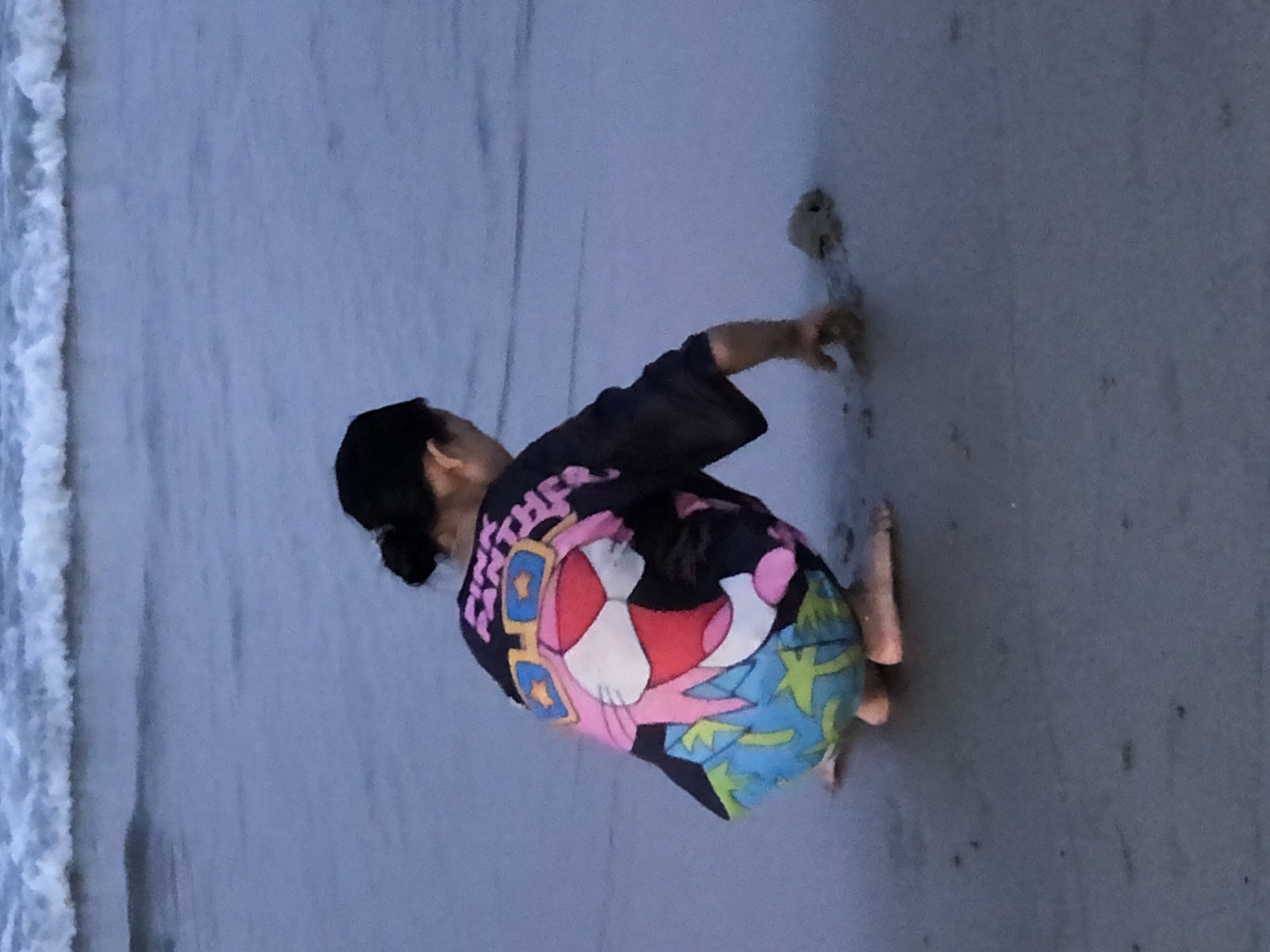 A person is seen from behind, crouching on a beach with wet sand and the edge of the ocean. The person is wearing a dark t-shirt with a colorful graphic on the back, featuring a cartoon character with pink fur, large blue sunglasses, and a patterned background of blue and green shapes. The character appears to be a stylized cat or similar animal. The person's right hand is reaching out towards the wet sand, seemingly interacting with something or simply touching the ground. Bare feet are visible, suggesting the person is barefoot.
The scene is set on a beach, with the gentle waves of the ocean visible on the left side of the frame, creating a foamy white line where the water meets the sand. The sand appears damp and dark, indicative of recent wave activity. The lighting suggests it might be dusk or dawn, as the overall tone is muted and the light is soft. There are some small dark spots on the sand, possibly debris or small pebbles.
The image appears to be taken in Đà Nẵng, Vietnam, a coastal city known for its beaches. No specific event is clearly depicted, but the activity suggests a moment of quiet observation or play on the beach. The person's posture and the focus on interacting with the sand could imply a sense of curiosity or engagement with the natural environment. The overall mood is calm and peaceful. No legible text is visible within the image itself, other than the stylized text on the t-shirt graphic which is partially obscured and difficult to read.