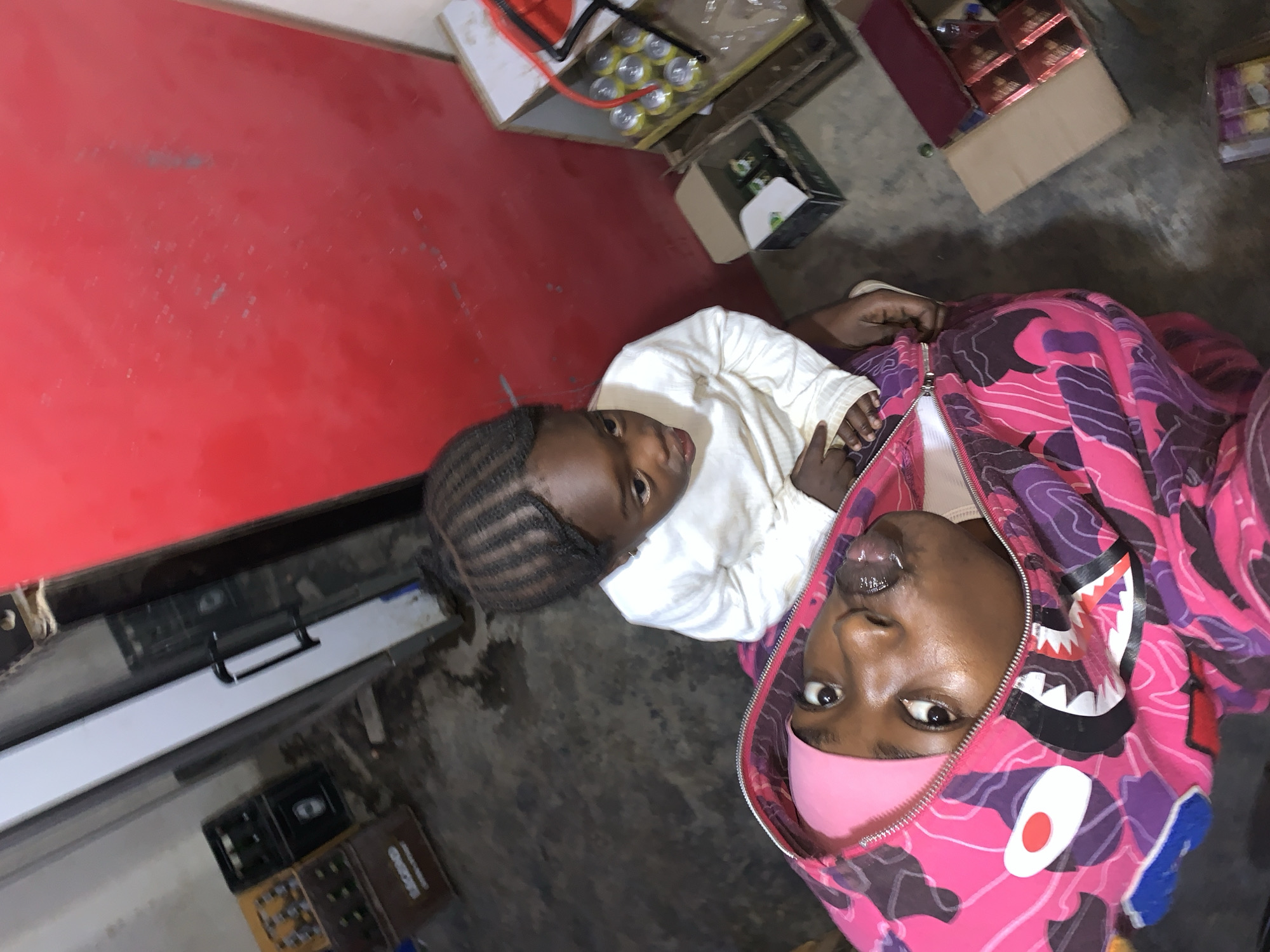 The image appears to be taken indoors in what may be a shop or storage area near New Stadium, Nigeria. Two individuals are visible. A child with braided hair is wearing a white shirt. Beside the child, an adult is wearing a pink and purple camouflage hoodie with a shark design on the hood and a pink mask covering their lower face. Around them are several boxes and containers, including beverages. A red surface is also visible to the left. The image is taken from an overhead angle.
