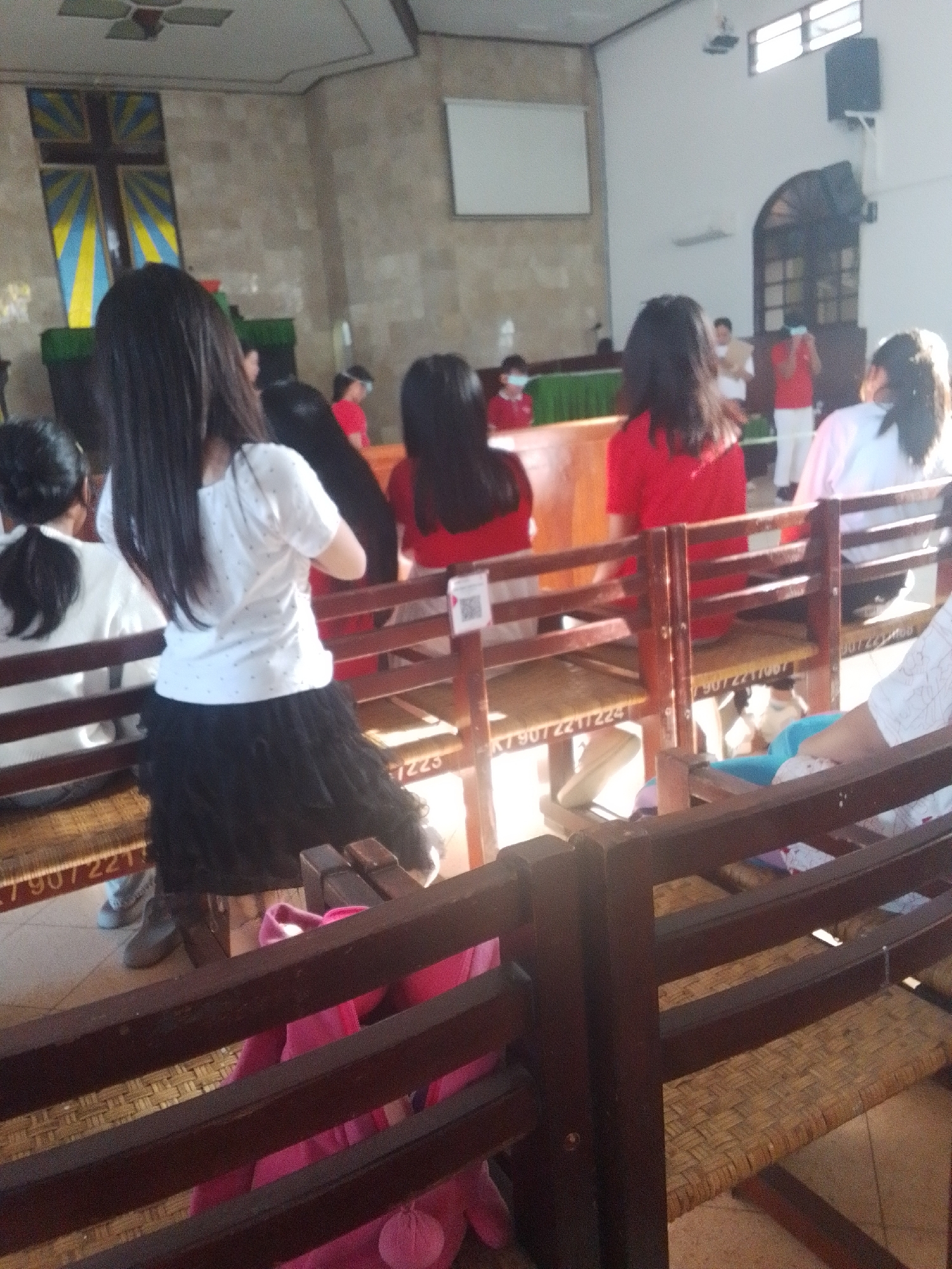 The image shows a group of people, mostly young women and girls, seated in wooden pews inside what appears to be a church or a place of worship. The interior features a large cross hanging on the back wall, which is adorned with a patterned fabric. The pews are made of dark wood with woven seating. Several individuals are wearing light-colored tops, and some are in red or pink. One girl in the foreground is wearing a white t-shirt and a black, ruffled skirt. The lighting suggests an indoor setting with natural light coming from windows. The overall atmosphere seems to be one of a religious gathering or service. The image does not provide explicit details that would allow for a definitive location in Makassar, Indonesia.