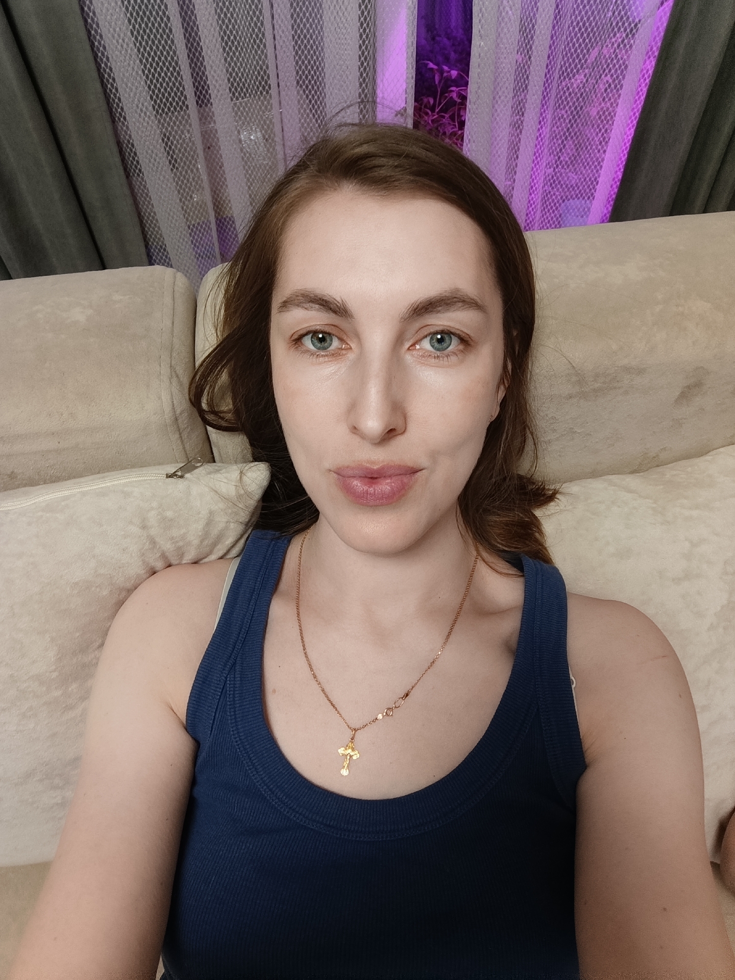 A young woman with light skin, long brown hair, and blue-green eyes is centered in a portrait-style image. She looks directly at the camera with a neutral, calm expression, her lips slightly parted. She is wearing a dark blue ribbed tank top and a gold chain necklace with a small gold crucifix pendant. The woman is seated or reclining on a light beige upholstered sofa, with matching cushions providing a soft backdrop. In the background, dark grey curtains are partially visible, overlaid by sheer white mesh curtains. Behind the curtains, a distinct purple light illuminates what appears to be foliage or plants outside a window, suggesting an evening or nighttime setting indoors. The scene depicts a casual indoor environment, likely a residential space in Chaikovsky, Russia. There is no visible text in the image.