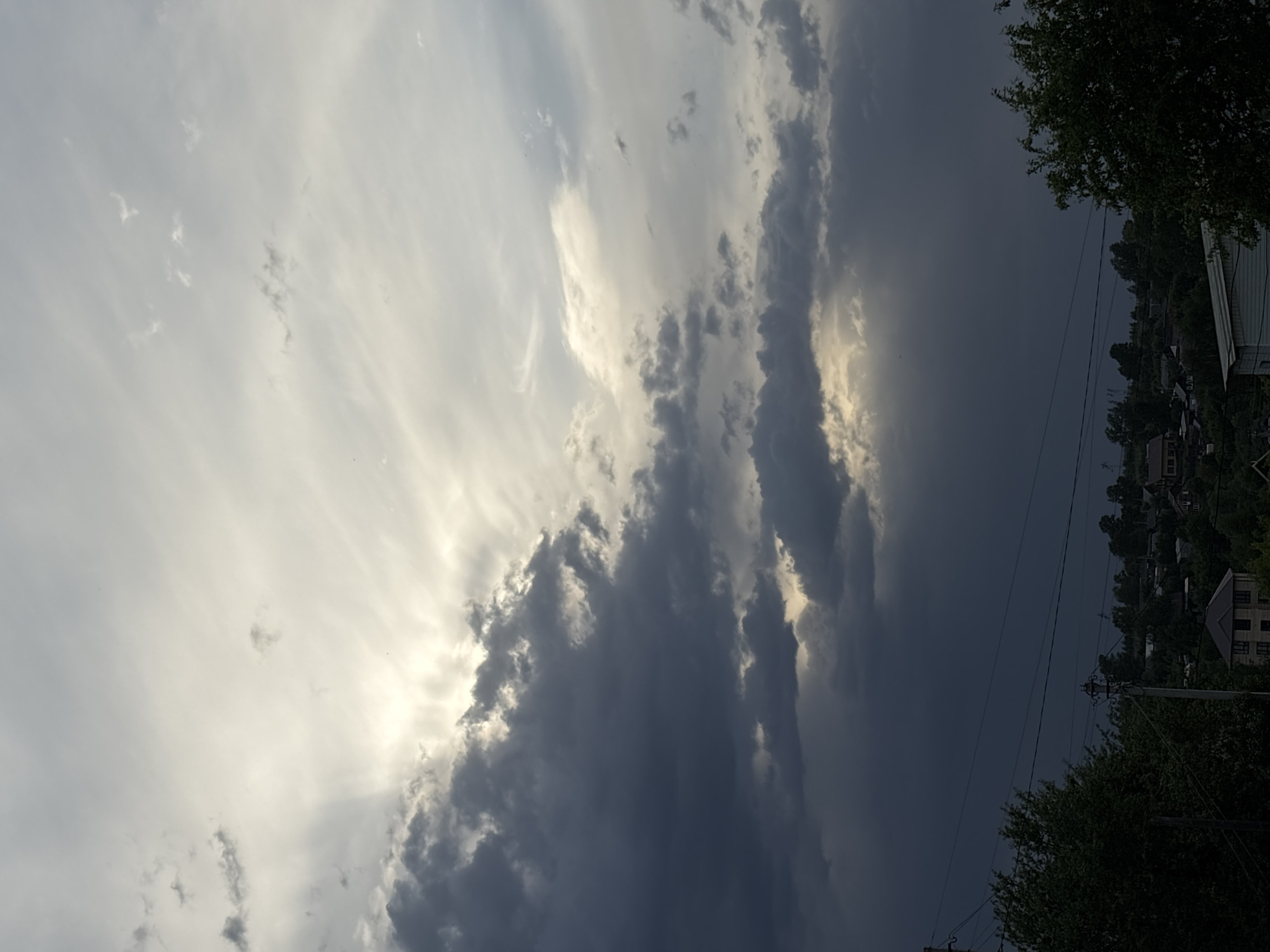 The image shows a dramatic sky with dark storm clouds contrasted against brighter, sunlit clouds. In the lower right of the frame, residential buildings and lush greenery are visible, suggesting a populated area. Electrical wires are strung across the foreground, a typical feature of urban or suburban environments. Based on the location Фоки, Russia, the scene is likely a residential area within the city, experiencing unsettled weather conditions.