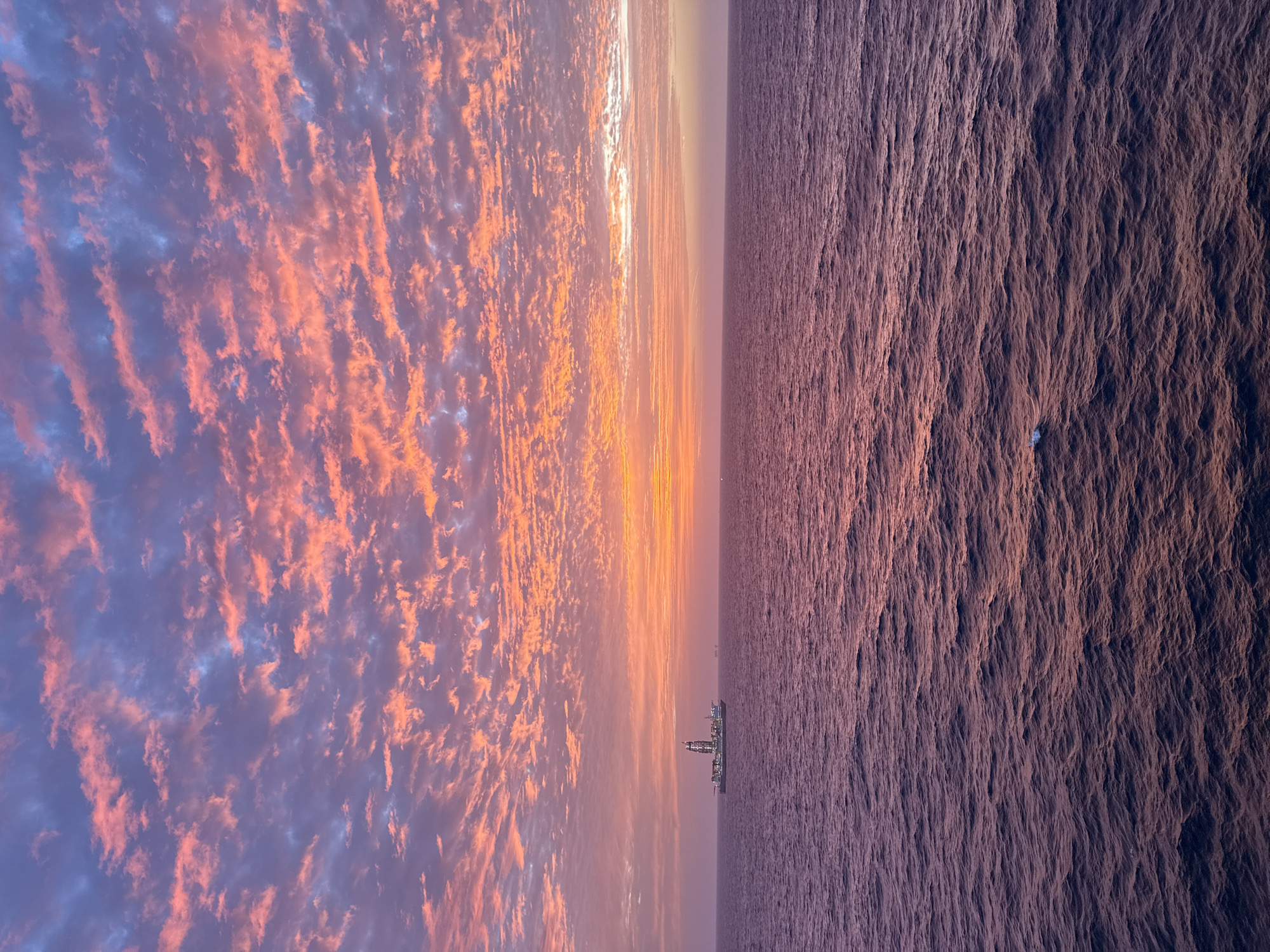 The image depicts a sky with textured clouds on the left, transitioning to a body of water on the right. A horizontal band of lighter sky separates the two elements. A structure, possibly a building or oil rig, is visible on the horizon between the sky and water. The scene is bathed in shades of pink, orange, and purple, suggesting a sunrise or sunset. The location is unknown.