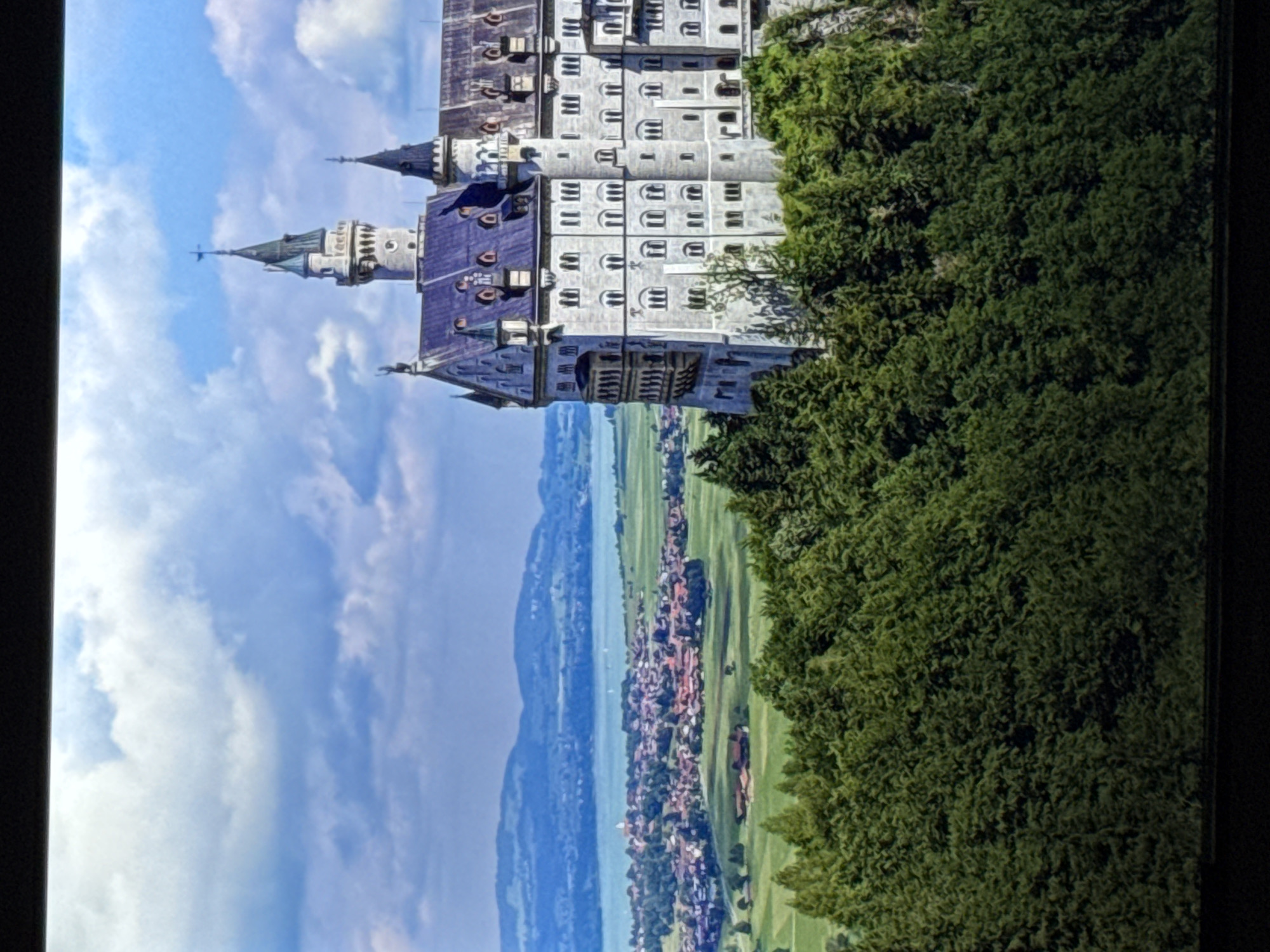 The image displays a majestic castle perched atop a lush green hill, overlooking a picturesque landscape. The castle, with its grand architecture, pointed turrets, and numerous windows, dominates the right side of the frame. Below the castle, a village with red-tiled roofs is nestled amidst rolling green fields. In the distance, a body of water, possibly a lake or a large river, stretches towards a range of hazy blue mountains. The sky above is a canvas of white, fluffy clouds against a bright blue backdrop, suggesting a pleasant day. The foreground is filled with dense evergreen trees, creating a natural border for the scene. There are no people visible in the image. This image does not depict any location in the city of Saratov, Russia.