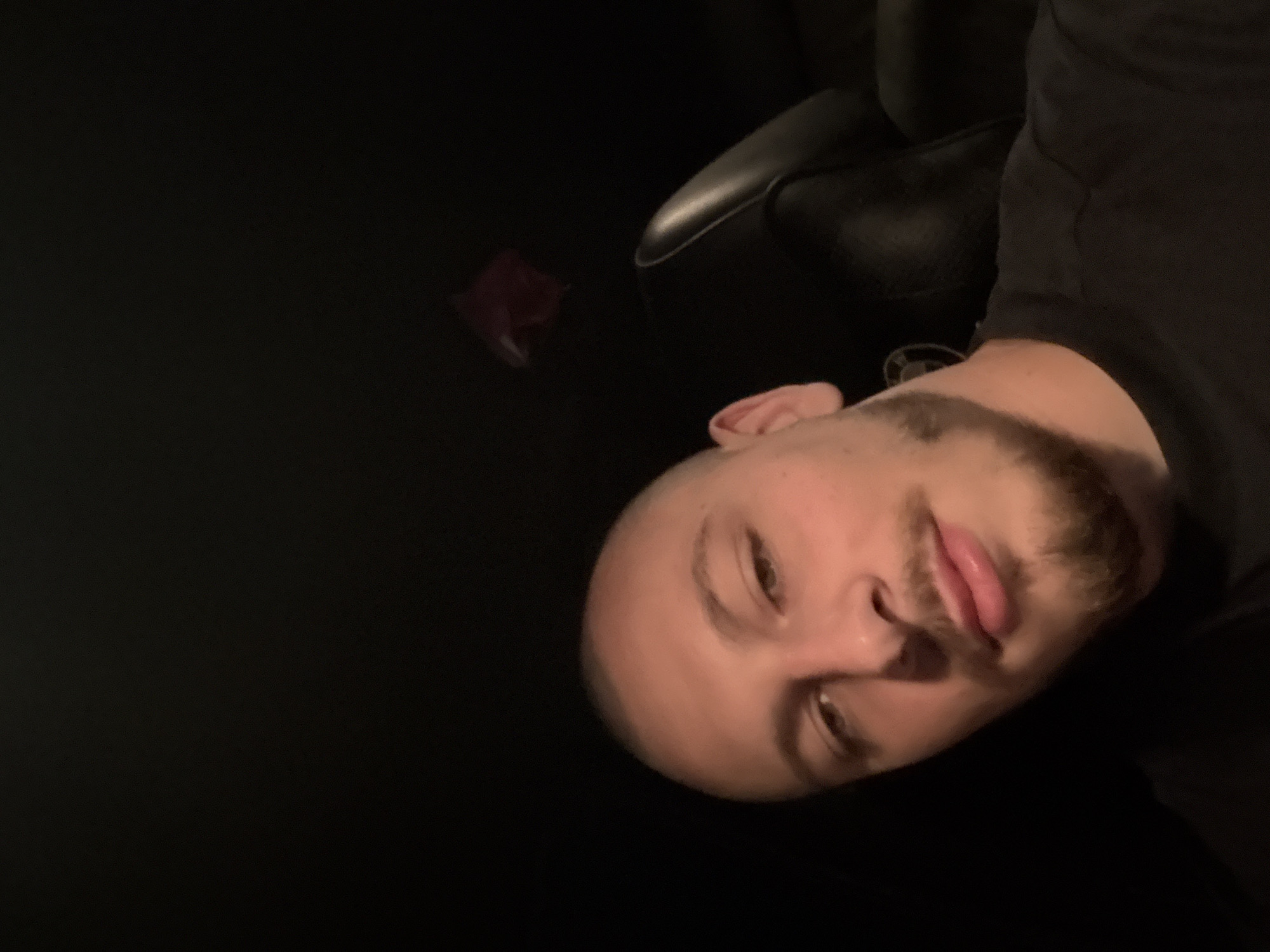 The image shows a close-up, low-angle selfie of a bald, light-skinned man with a goatee and mustache, wearing a black t-shirt. He is looking at the camera with a neutral expression. The background is dark, with a hint of a dark leather seat and a small, blurry red object in the upper right corner, possibly a car accessory or decoration. The overall impression is that the photo was taken inside a vehicle at night. There is no information in the image that allows for a determination of the specific location in Stara Zagora, Bulgaria.