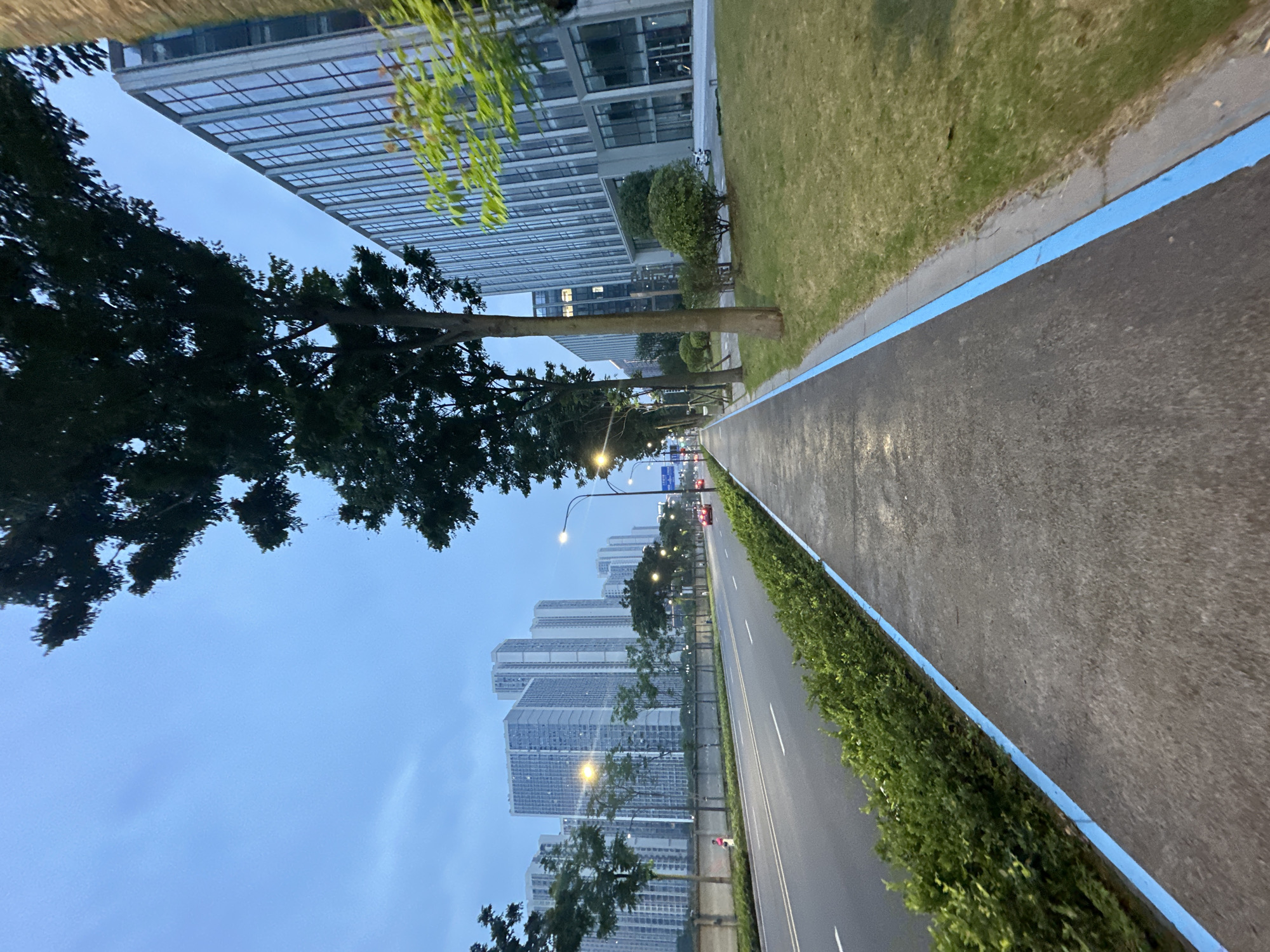 The image depicts a sidewalk running alongside a road in the Chongqing High-tech Zone, China, during twilight. Tall, modern buildings with glass facades line the right side of the road, while a row of apartment buildings is visible in the distance on the left. Trees and manicured green hedges border the sidewalk and the road. The sky is overcast and a pale blue. The sidewalk has a distinct blue line painted along its edge. A few cars are on the road, and a single person is walking on the sidewalk in the distance. The scene appears quiet and orderly, typical of an urban environment during the evening transition.