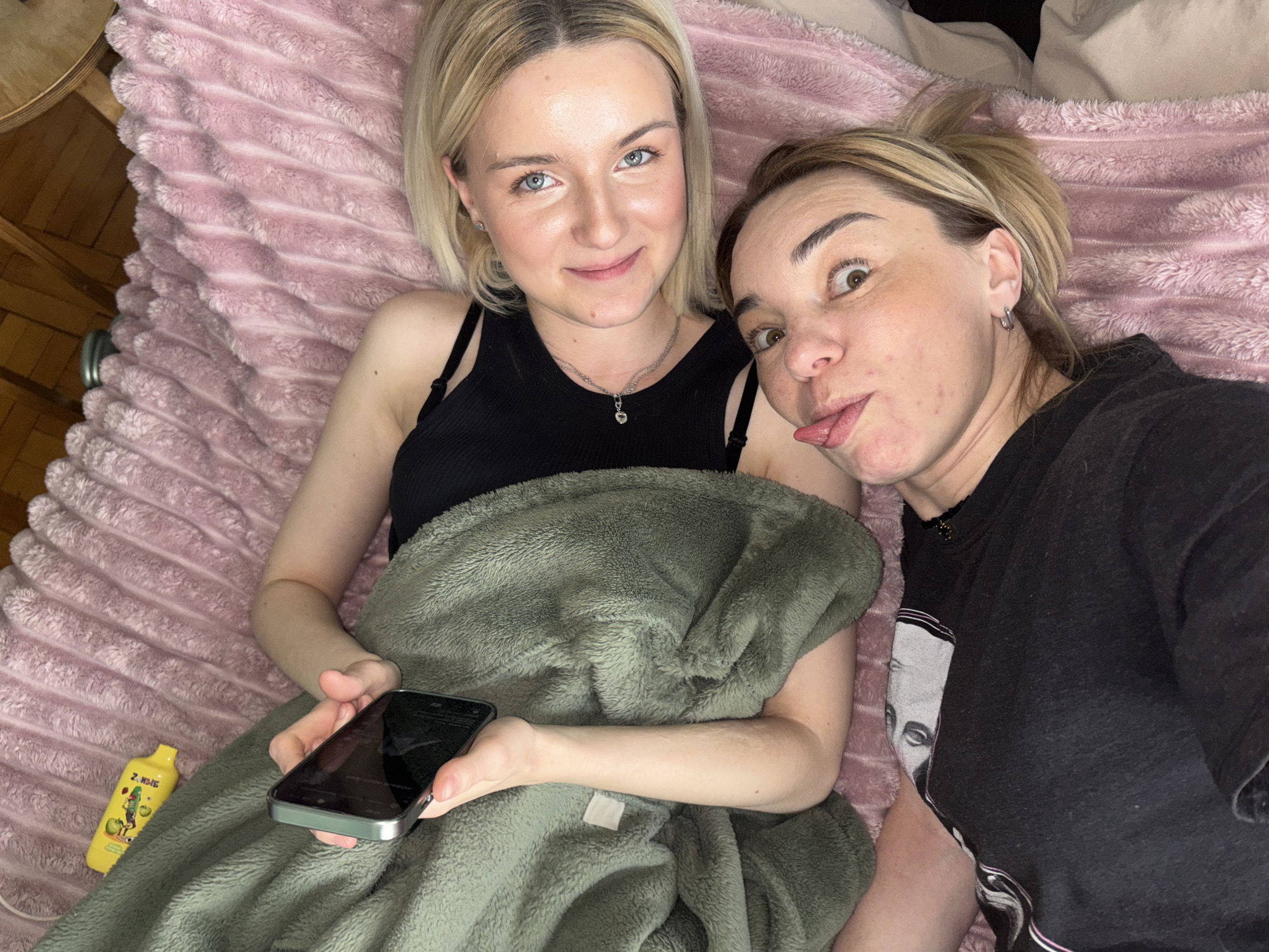 Two women are lying down indoors, likely on a bed or sofa, captured from an overhead perspective. A light pink, ridged blanket covers the surface they are on.
The woman on the left has short blonde hair and light blue eyes, looking directly at the camera with a gentle smile. She is wearing a black tank top and holds a smartphone in her left hand. Her lower body is covered by a green fuzzy blanket. A small silver heart pendant hangs from a necklace around her neck.
The woman on the right has blonde and light brown hair pulled back, with brown eyes. She is making a playful, humorous face, sticking out her tongue and widening her eyes. She wears a black t-shirt, part of which features a faded graphic design that is not fully discernible but appears to include a face. She has an earring in her visible ear.
To the left of the blonde woman, near her hip, rests a yellow vape pen. Visible text on the vape pen clearly reads "ZOMBIE" alongside an image of green apples. The setting appears to be a cozy indoor environment, possibly in Vsevolozhsk, Russia, with wooden flooring visible at the top left edge. The overall mood is relaxed and lighthearted, suggesting a moment of casual companionship or a selfie taken during downtime.