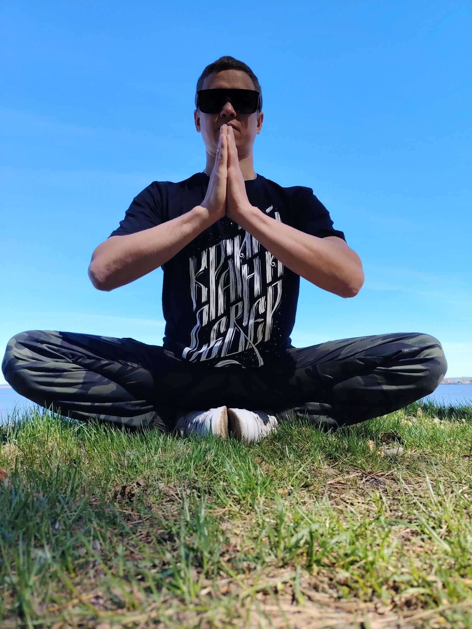 A man is seated in a meditative pose outdoors. He is wearing a black t-shirt with a white graphic design, camouflage pants, and sunglasses. His hands are pressed together in front of his face. He appears to be sitting in the lotus position on grass. The sky is clear and blue. A body of water and a distant shoreline are visible in the background.
While the location is asserted to be Chaikovsky, Russia, there is no information in the image to independently confirm this statement.