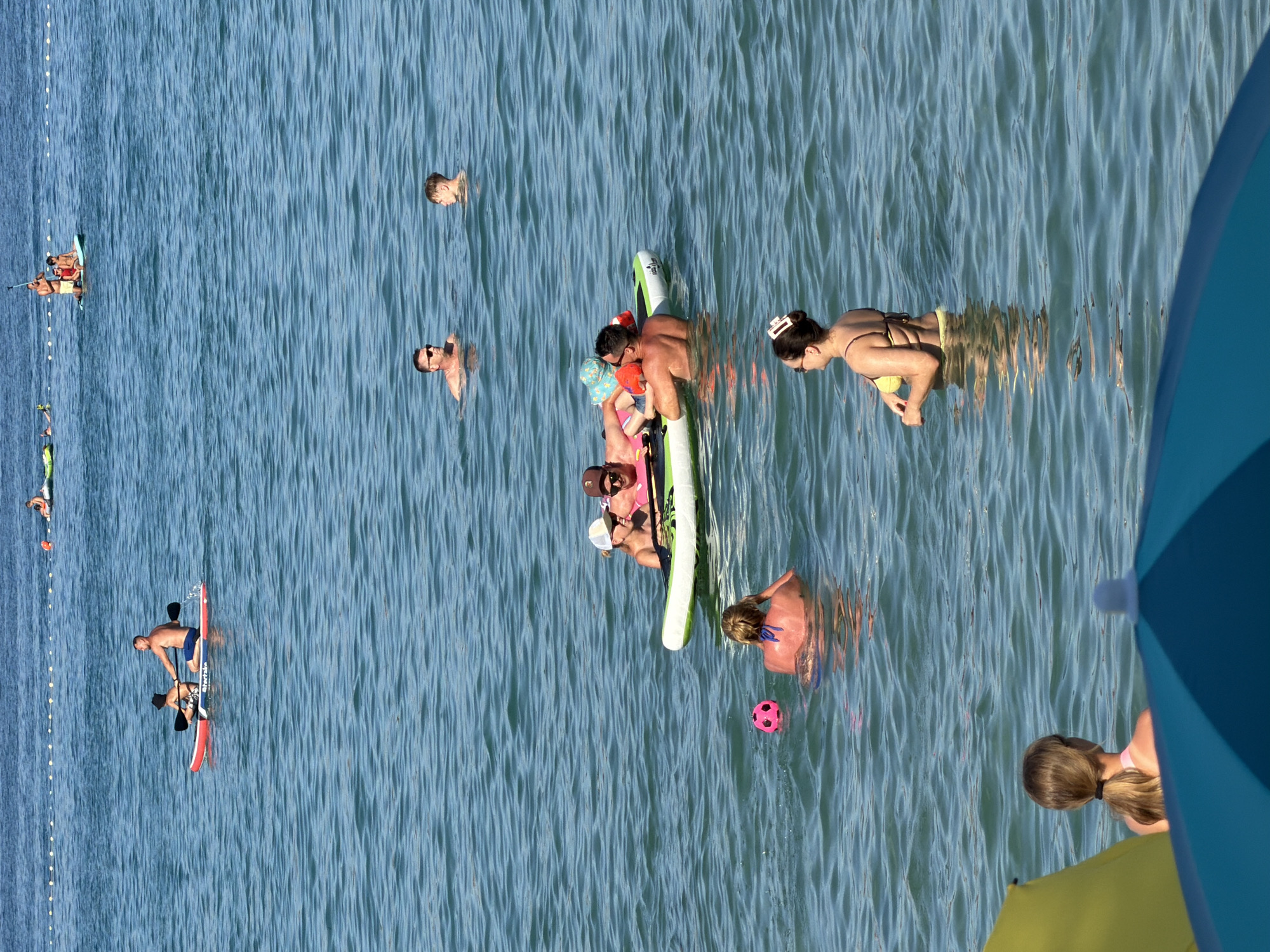 The image shows a sunny day at sea, with several people engaged in water activities. In the foreground, a yellow umbrella and a blue umbrella partially obscure the view of a woman facing away from the camera, with her hair tied in a ponytail, wearing a pink top. Further out, a woman in a yellow bikini is standing in the water. In the middle ground, a group of four people are on an inflatable raft, with two children and two adults. A pink ball floats in the water nearby. Further out, a man and a child are paddleboarding, with the man paddling. In the background, several other individuals are visible in the water and on paddleboards. A rope with buoys stretches across the water in the far background. The water is blue and calm with ripples. The location is identified as the sea in Općina Baška, Croatia.