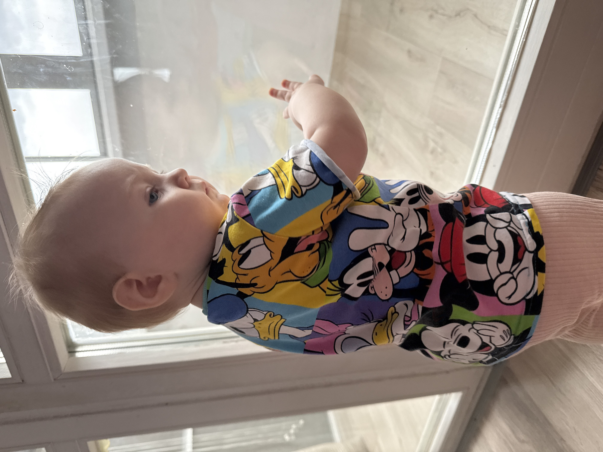 The image, oriented sideways, features a baby primarily in profile, looking out of a large window. The baby has fair skin, light blonde hair, and blue eyes, and appears deeply curious and engaged with the view outside.
The baby is wearing a brightly colored t-shirt adorned with various cartoon characters, including recognizable figures like Mickey Mouse, Donald Duck, and Goofy, depicted in a pop-art style with bold outlines and vibrant colors such as blue, yellow, red, green, and pink. They are also wearing light-colored, possibly pink or peach, ribbed pants. One of the baby's hands is slightly raised, appearing to rest against or reach towards the window glass.
The setting is indoors, likely a home in Chaikovsky, Russia, with a light-colored wooden or laminate floor visible towards the bottom and right of the frame. A white window frame outlines the transparent glass, through which diffused daylight streams, suggesting it is daytime, possibly with an overcast sky. No specific objects are clearly discernible outside the window, only a bright, uniform light. There is no visible text in the image.
