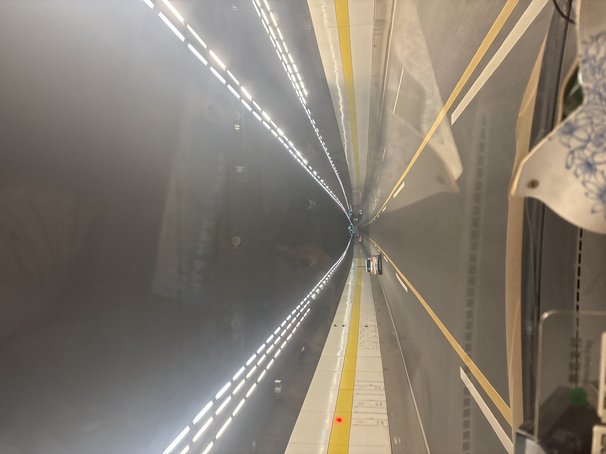 The image shows the interior of a car traveling through a tunnel. The tunnel has a dark ceiling with rows of bright white lights on either side. The walls of the tunnel are white with a horizontal yellow stripe. The road is asphalt with yellow and white lane markings. There are several cars visible ahead in the tunnel. In the foreground, part of the car's dashboard and windshield are visible, along with a tissue box with a floral pattern. The image is taken from the perspective of the driver. Based on the provided context, this scene is located in Xiangcheng District, China.