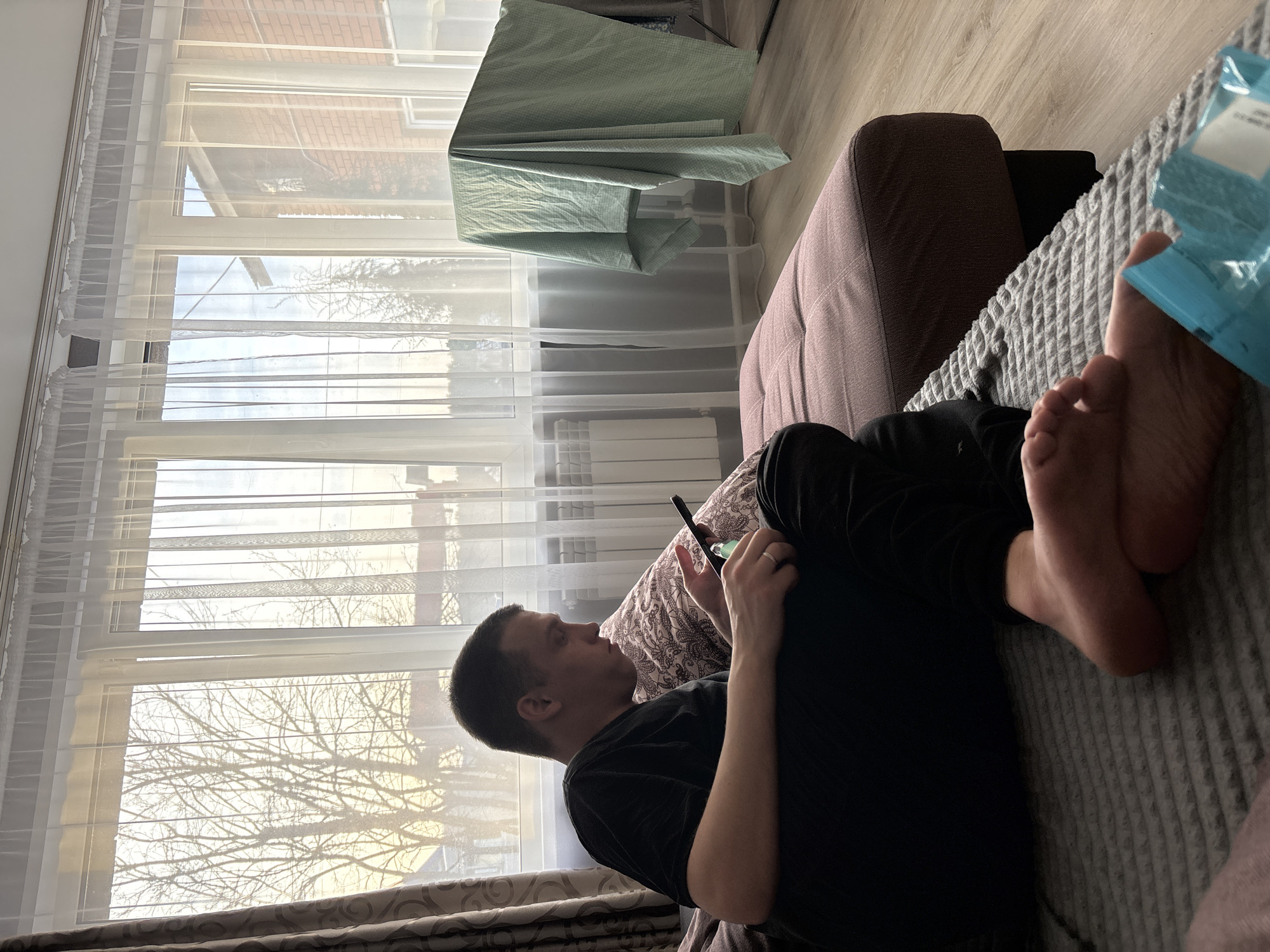 The image depicts an indoor scene in Chaikovsky, Russia, looking out through large windows. A male individual, with short dark hair, is seated on a brown sofa, leaning against patterned pillows. He wears a black t-shirt and black pants, with his bare feet resting on a gray textured blanket. He is actively engaged with a black smartphone, holding it with both hands and looking down at its screen. A silver-colored ring is visible on his left ring finger.
The room features a light wood-patterned laminate floor. To the left, a portion of a dark gray or off-white wall is visible, with a white radiator mounted below the window. The windows themselves are multi-pane with white frames and are covered by translucent white sheer curtains.
Outside the window, bare trees are visible, suggesting a dormant season. The sky appears clear with soft sunlight. In the distance, structures including a building with reddish-brown brickwork and a white fence are discernible. To the right of the window, a light green checkered sheet or fabric is draped over a metallic clothes drying rack. A small, light blue plastic package lies on the gray blanket near the man's feet.