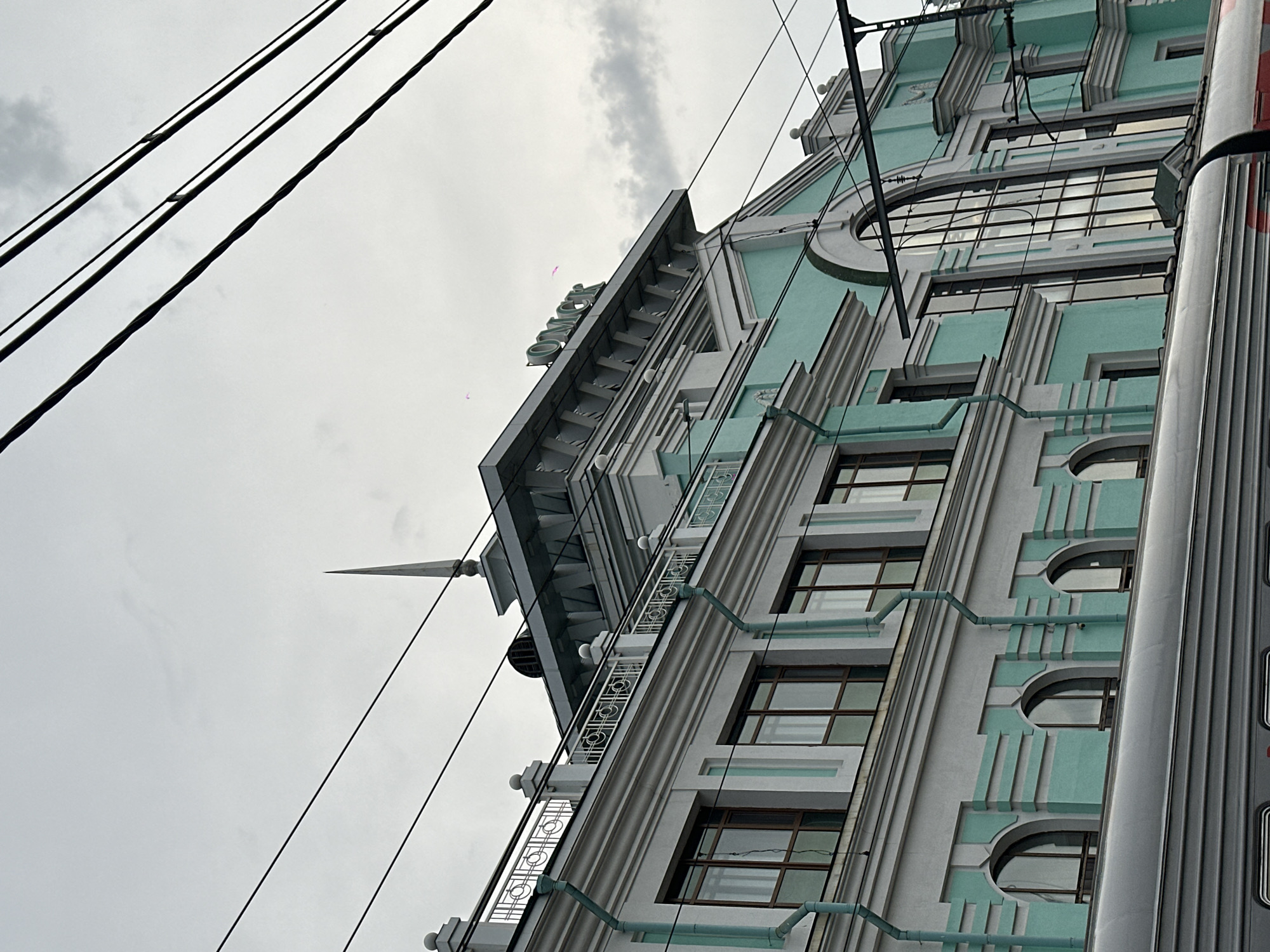 This is an exterior, eye-level, medium shot of the Omsk railway station in Omsk, Russia. The building is painted in a light gray and turquoise color scheme with ornate architectural details. A train car is visible in the foreground, partially obscuring the lower portion of the building. Overhead, multiple dark electrical wires crisscross the frame, silhouetted against a cloudy, overcast sky. The word "ОМСК" (Omsk) is prominently displayed in green lettering on the upper facade of the building. The architecture suggests a classical or neoclassical style. There are no people visible in the image. The scene is static and appears to be captured during daylight hours.
