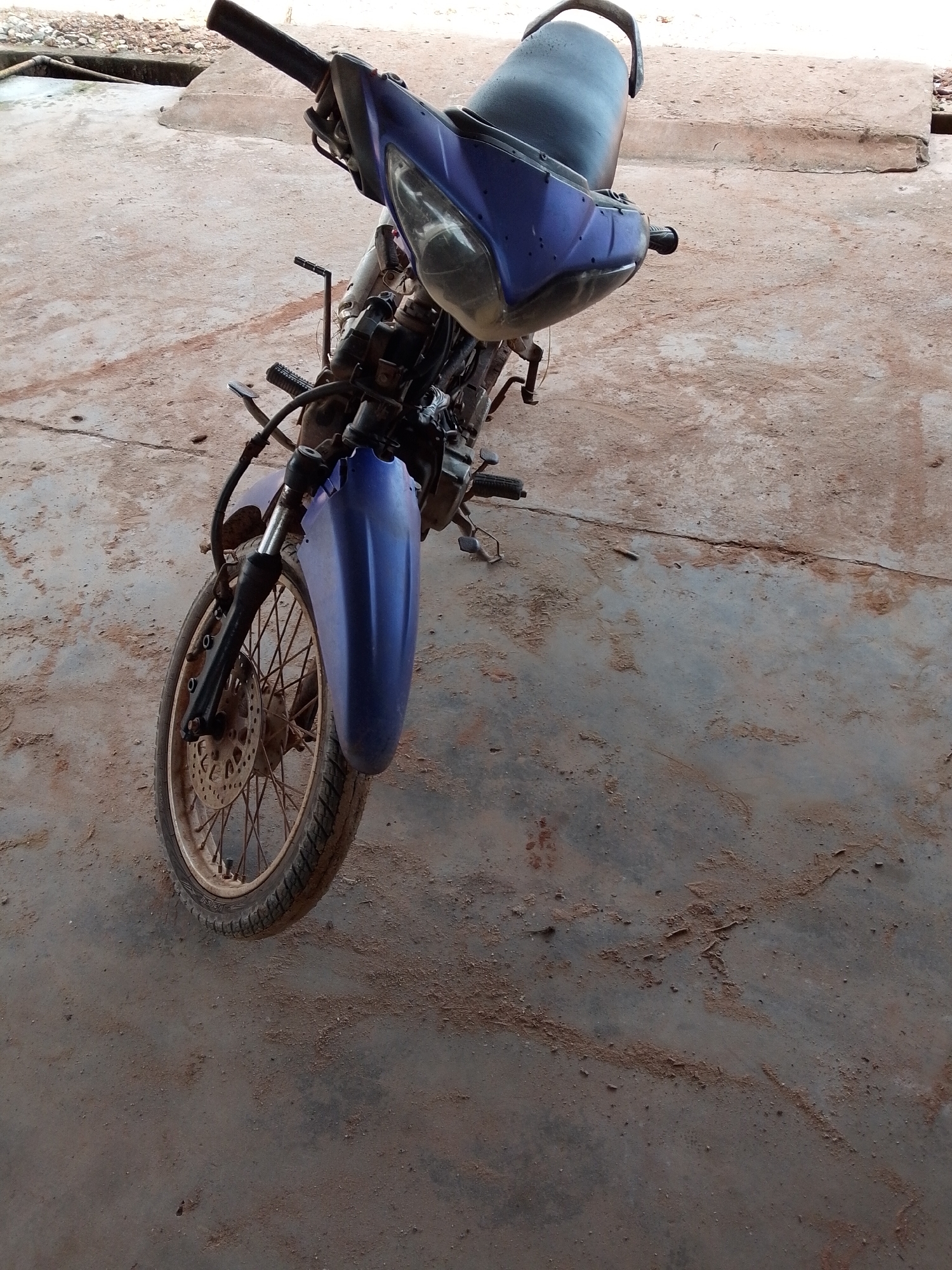 The image depicts a partially disassembled, blue motorcycle parked on a dirt-covered concrete surface. The motorcycle's front wheel is heavily soiled, and parts of the bike appear rusty or dirty. The surrounding environment is a weathered concrete area, suggesting an outdoor or garage setting. The location is Bukit Ketok, Indonesia. No people are visible in the image.