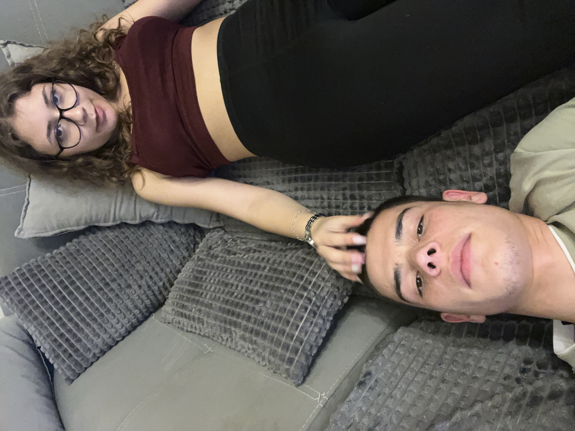Two individuals are lying on a gray couch with throw pillows. The first individual is a young female with curly brown hair and glasses, wearing a burgundy crop top and black leggings. She is positioned on the left, with her head resting on a gray pillow. The second individual is a young male, positioned on the right, wearing a light-colored shirt. The female's hand is on the male's head. The setting appears to be indoors, with the couch occupying most of the frame. There are no indicators of Plovdiv, Bulgaria.