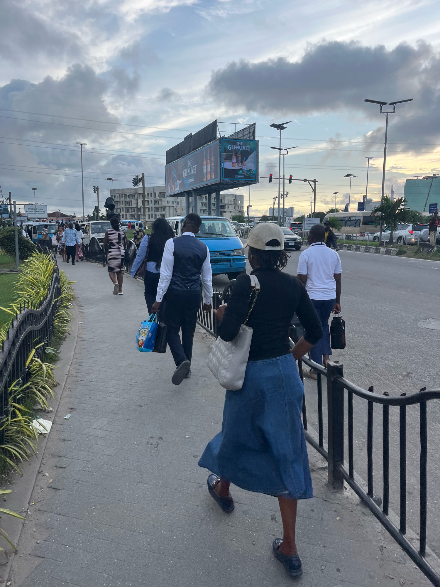 A medium shot captures people walking on a sidewalk in Lekki Phase I, Nigeria, near a busy road. The sky is overcast with dramatic clouds. On the left side of the sidewalk, lush green and yellow foliage lines a black metal fence. A man in a white shirt and dark pants walks away from the camera, carrying a black bag. Behind him, a woman in a denim skirt and black long-sleeved shirt, wearing a beige cap and carrying a white bag, also walks away. Further back, a group of people walk on the sidewalk, including a man in a dark vest over a white shirt and a woman in a patterned dress. On the road, a blue van and other vehicles are visible, along with a large billboard displaying "THE GLENLIVET". Streetlights and traffic signals are present in the background. The scene depicts an urban environment with moderate pedestrian and vehicle traffic.
