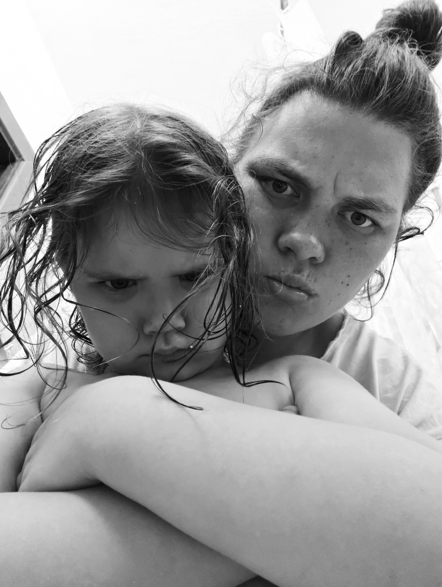 A black and white, close-up selfie-style photograph features two individuals: a child and an adult. Both subjects are facing the camera with stern expressions, pursed lips, and furrowed brows. The child, positioned in the foreground, appears to have wet hair. The adult is positioned behind the child. The adult has their hair pulled back in a bun. The location is Chaikovsky, Russia. The photograph captures a moment of perceived displeasure or resistance. The details suggest a domestic or personal setting.